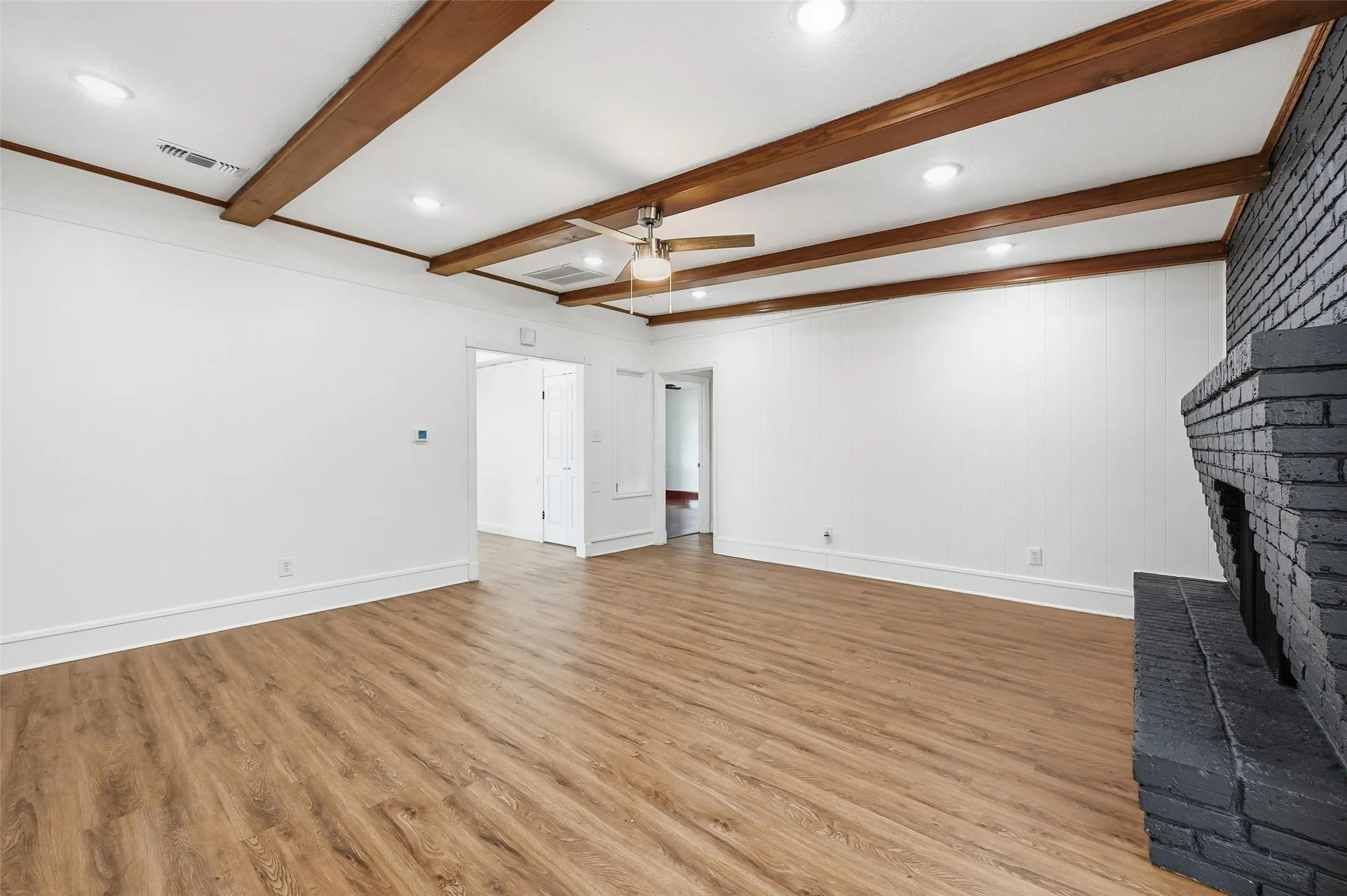 Spacious room featuring wood-finish flooring, a painted brick fireplace, exposed wood ceiling beams, recessed lighting, and a ceiling fan