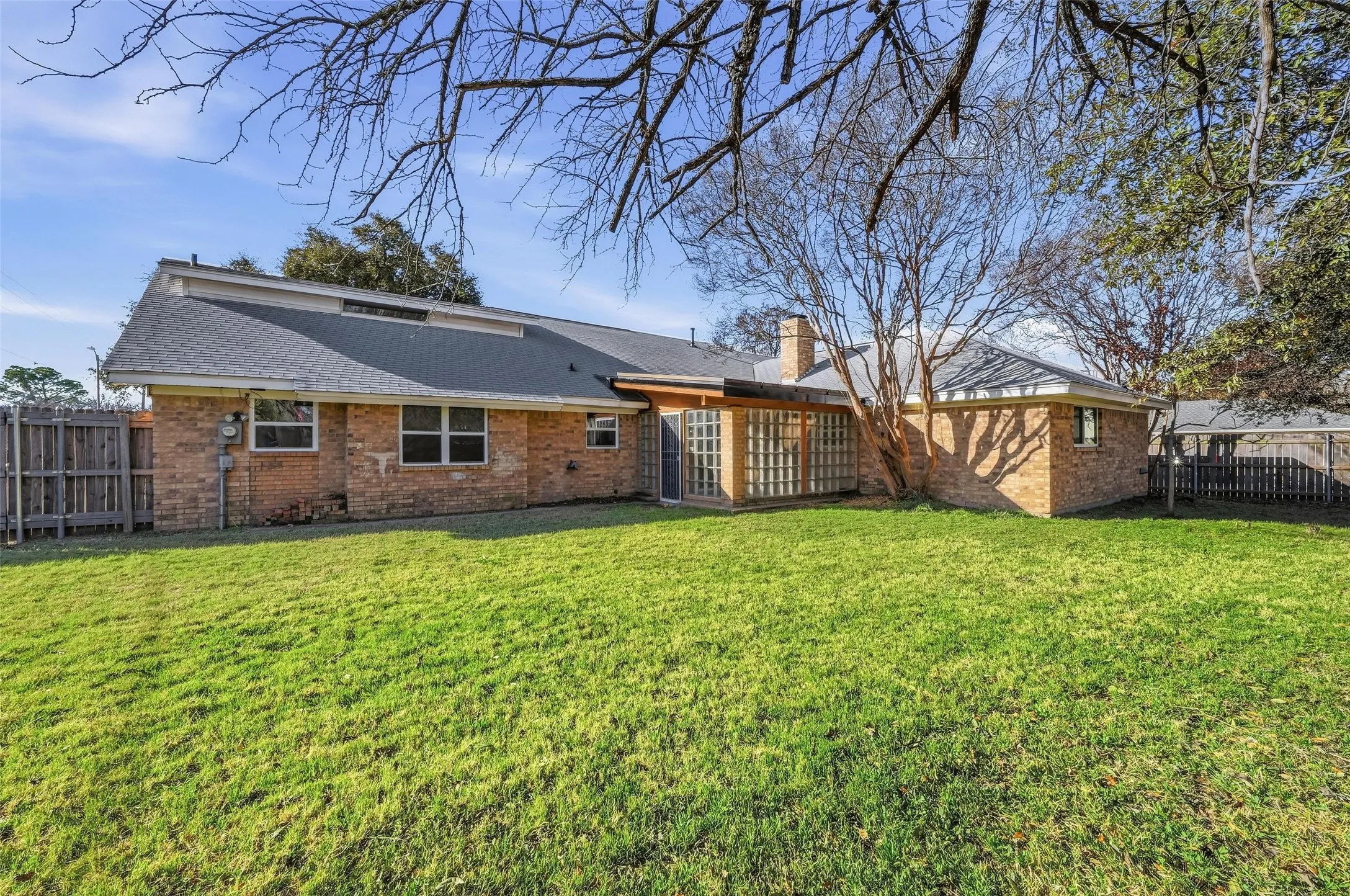 Spacious backyard with a brick exterior home, featuring a dark shingle roof, multiple windows, a wood privacy fence, and mature trees