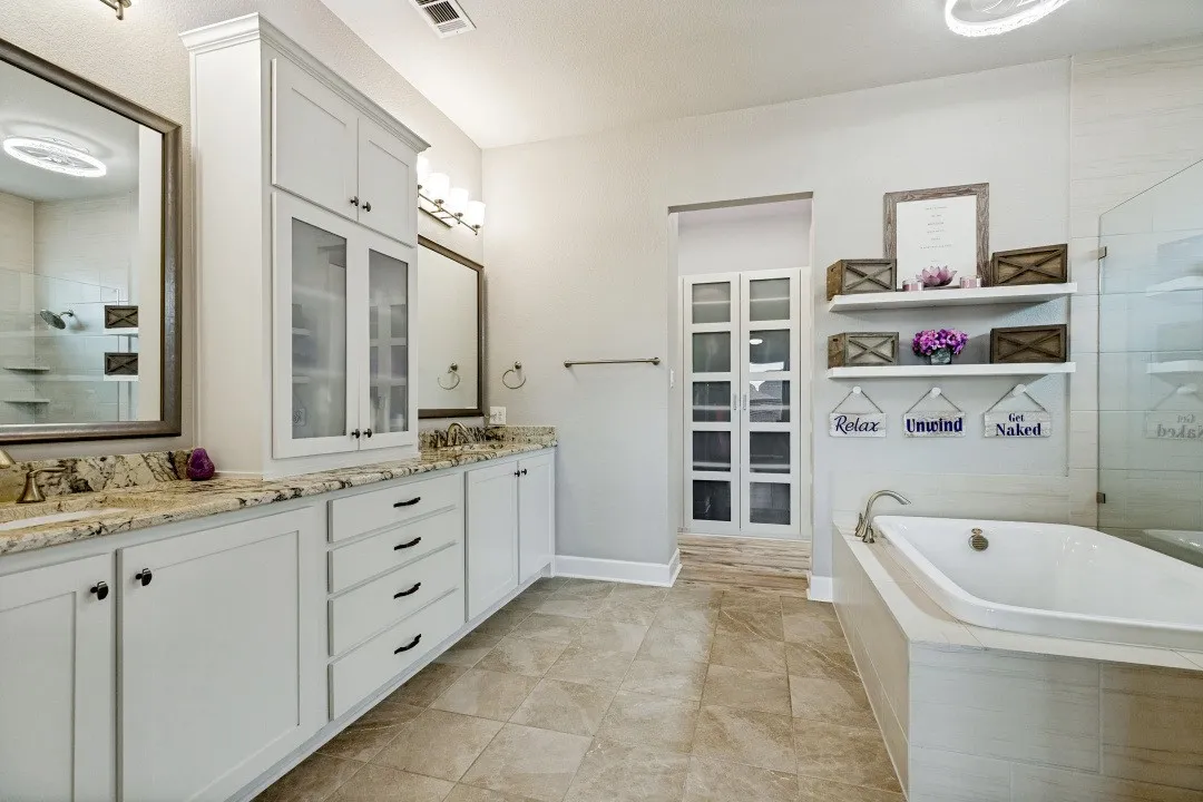 Spacious bathroom featuring dual vanities with light-toned countertops, a built-in soaking tub, a glass-enclosed shower, and ample white cabinetry including a tall linen cabinet with glass-paneled doors