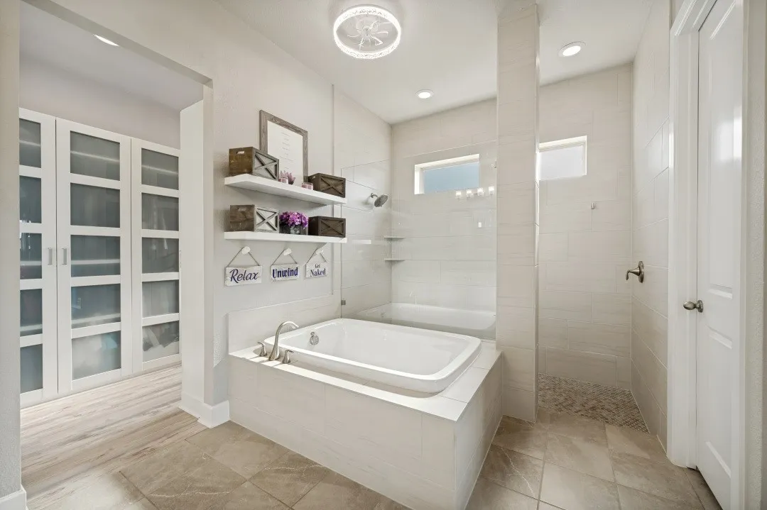 Spacious bathroom featuring a built-in soaking tub with brushed nickel fixtures, a walk-in shower with pebble flooring and tiled walls, a frosted glass privacy window, and a separate room with built-in cabinetry and glass-front doors