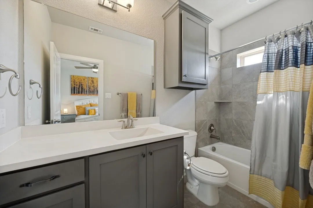 Bathroom featuring a vanity with a white countertop and gray cabinetry, a rectangular undermount sink, a large wall-mounted mirror, a tub/shower combination with gray tile surround, and a wall-mounted gray cabinet