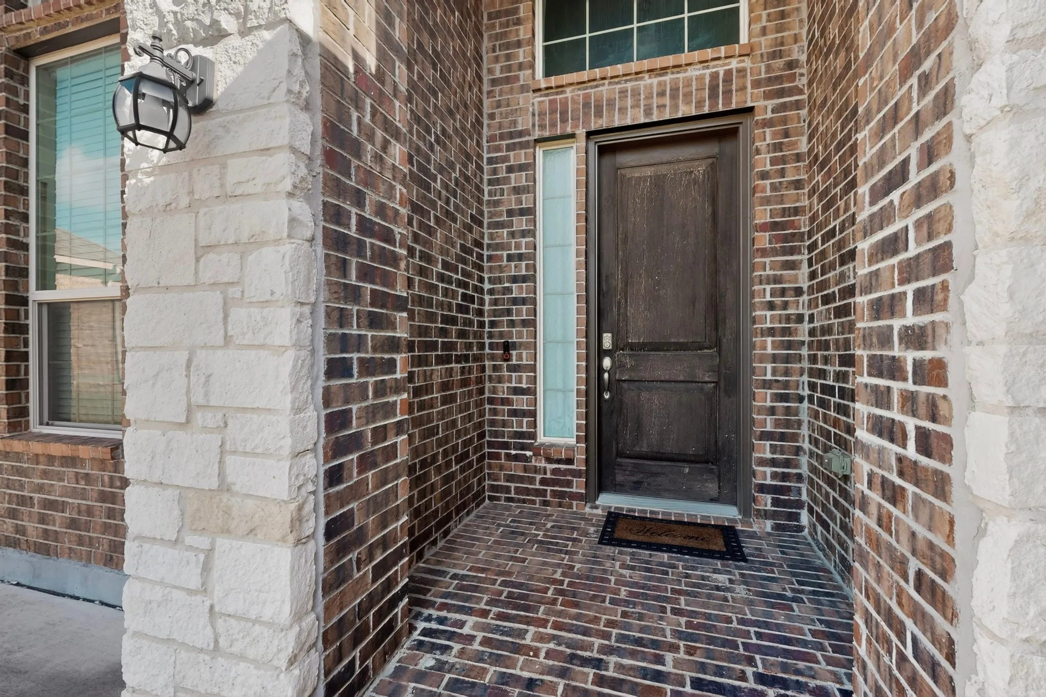 Entryway featuring a dark wood-finish door with sidelight, brick-patterned porch flooring, and a decorative outdoor lantern