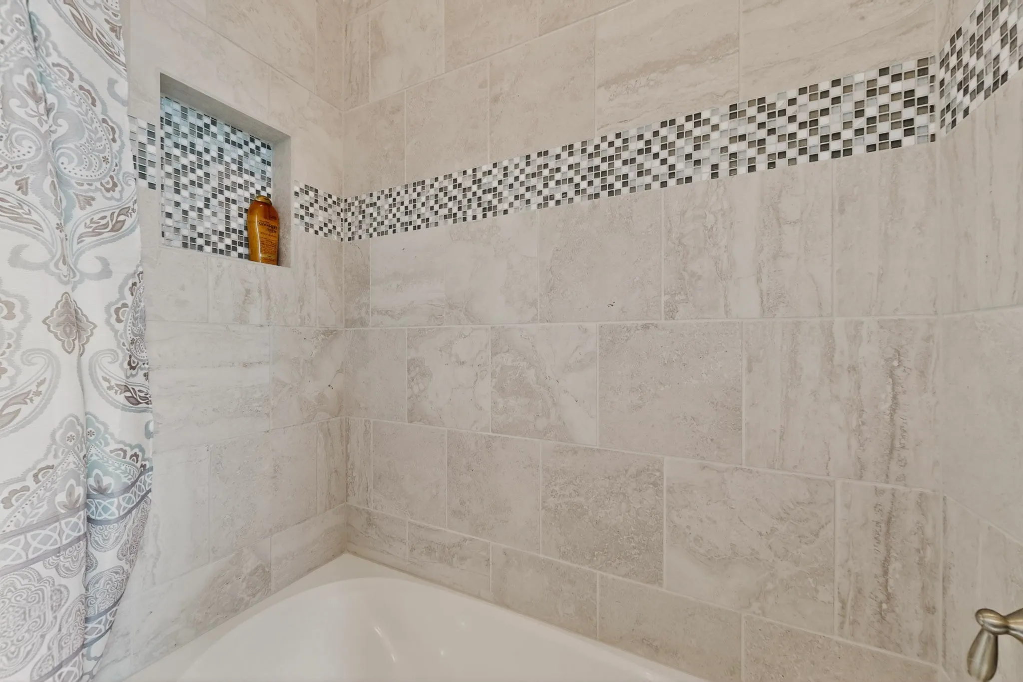 Bathtub with light-toned wall tile, a mosaic accent band, and a tiled shower niche