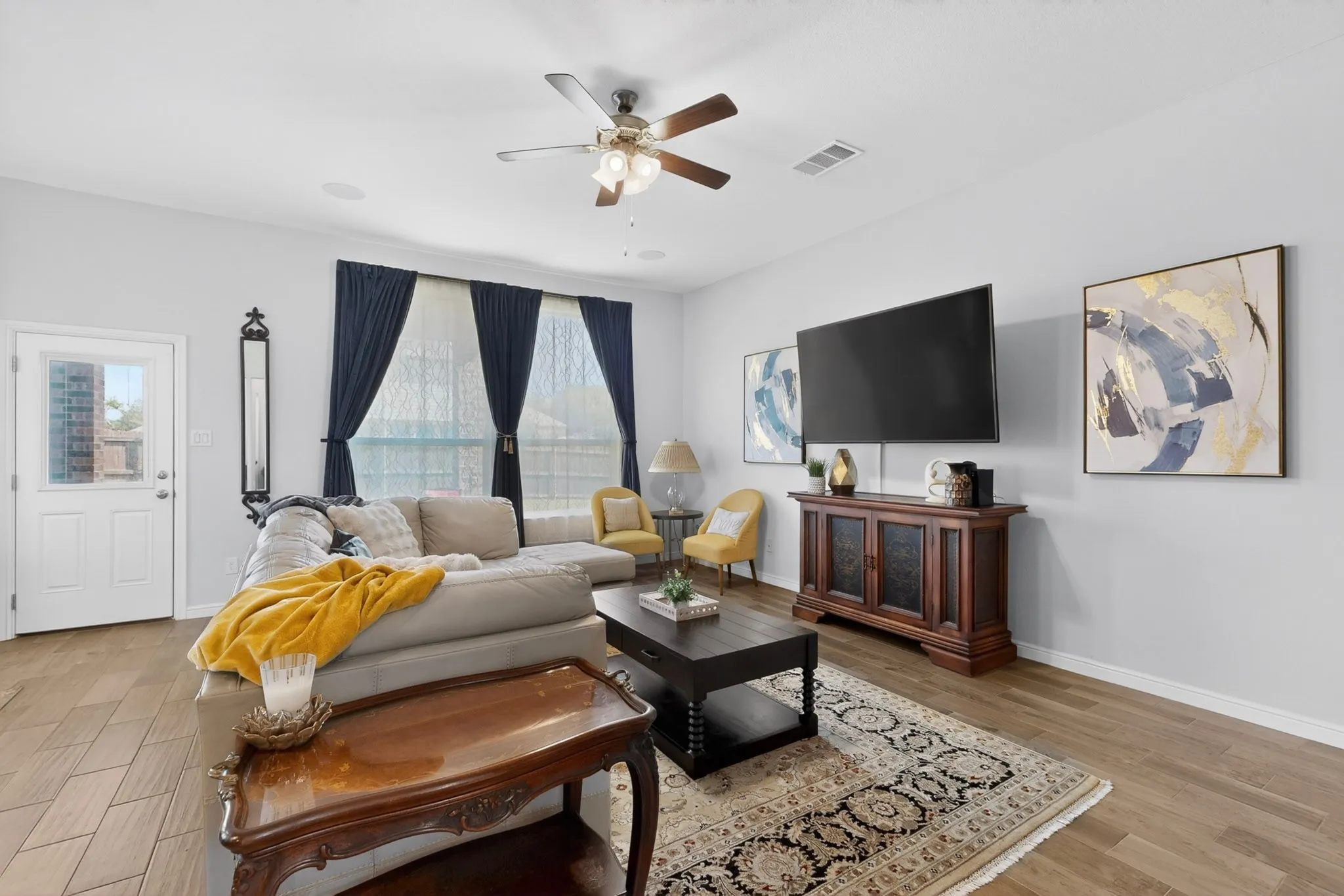 Spacious living area featuring wood-finish flooring, light gray walls, and large windows with privacy sheers