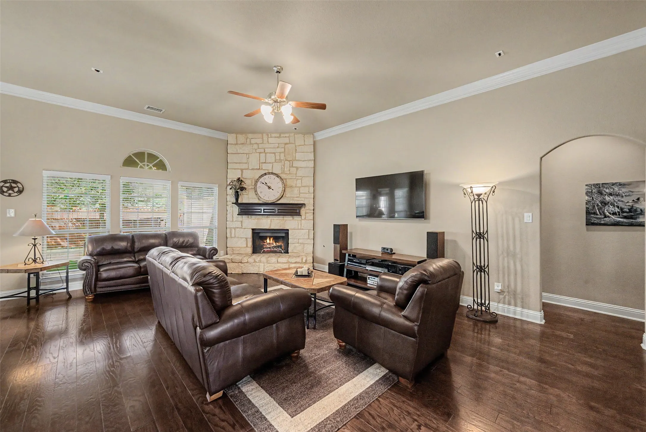Spacious living room featuring rich wood-finish flooring, a stone fireplace with a dark mantel, and multiple windows with an arched transom