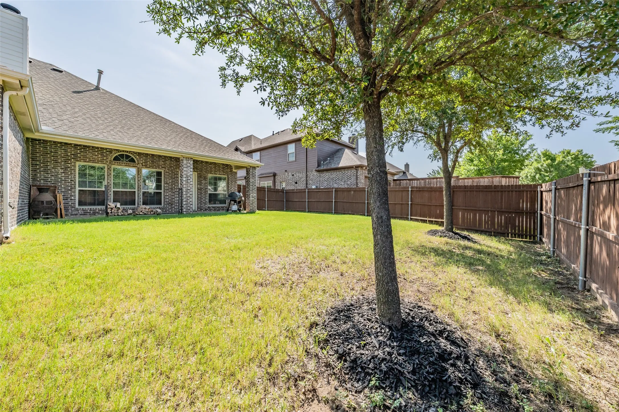 Spacious backyard featuring a brick exterior, covered patio, multiple mature trees, and privacy fencing