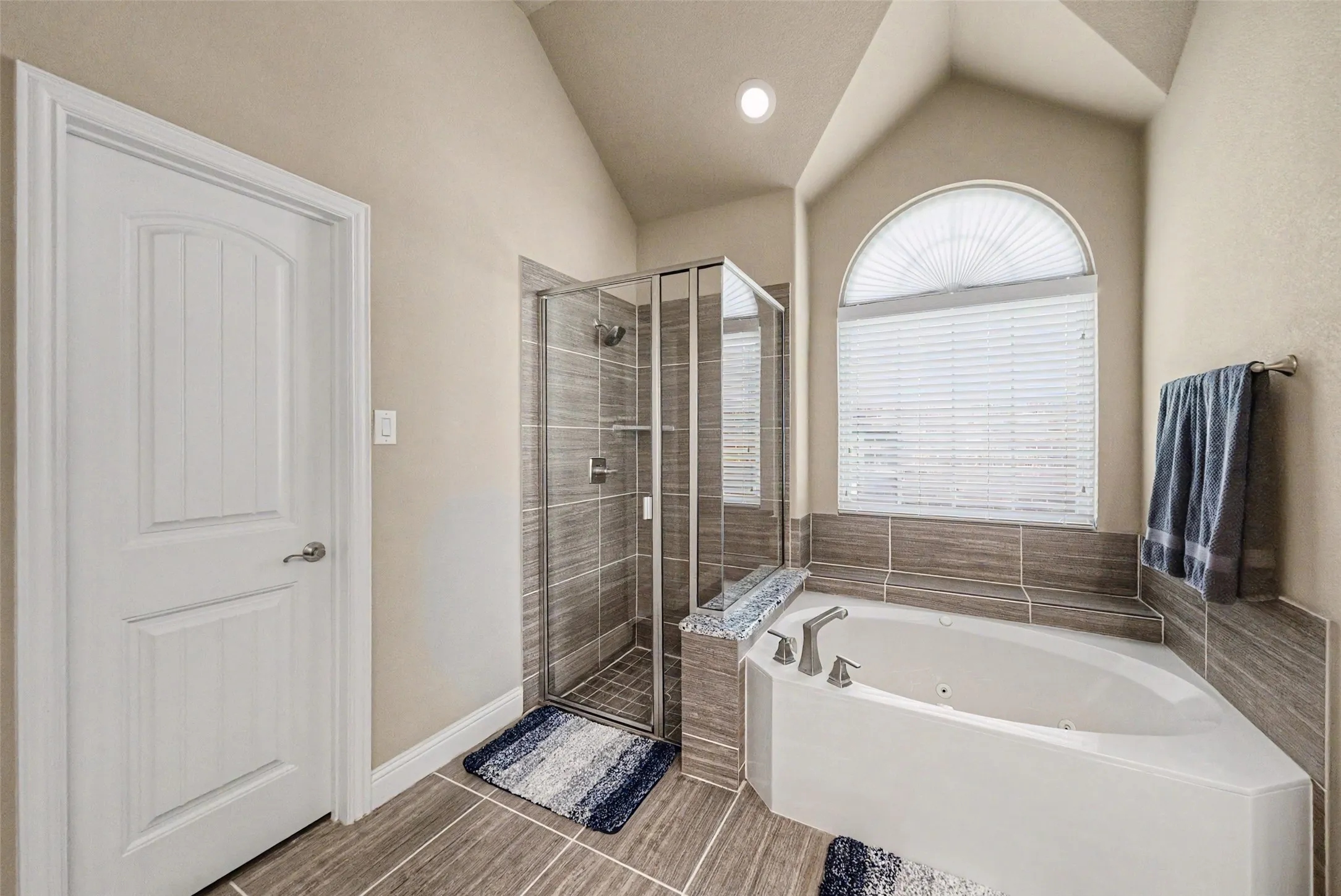 Bathroom featuring a corner soaking tub with deck-mounted fixtures, an arched window with horizontal blinds, and a tiled shower enclosure with a glass door