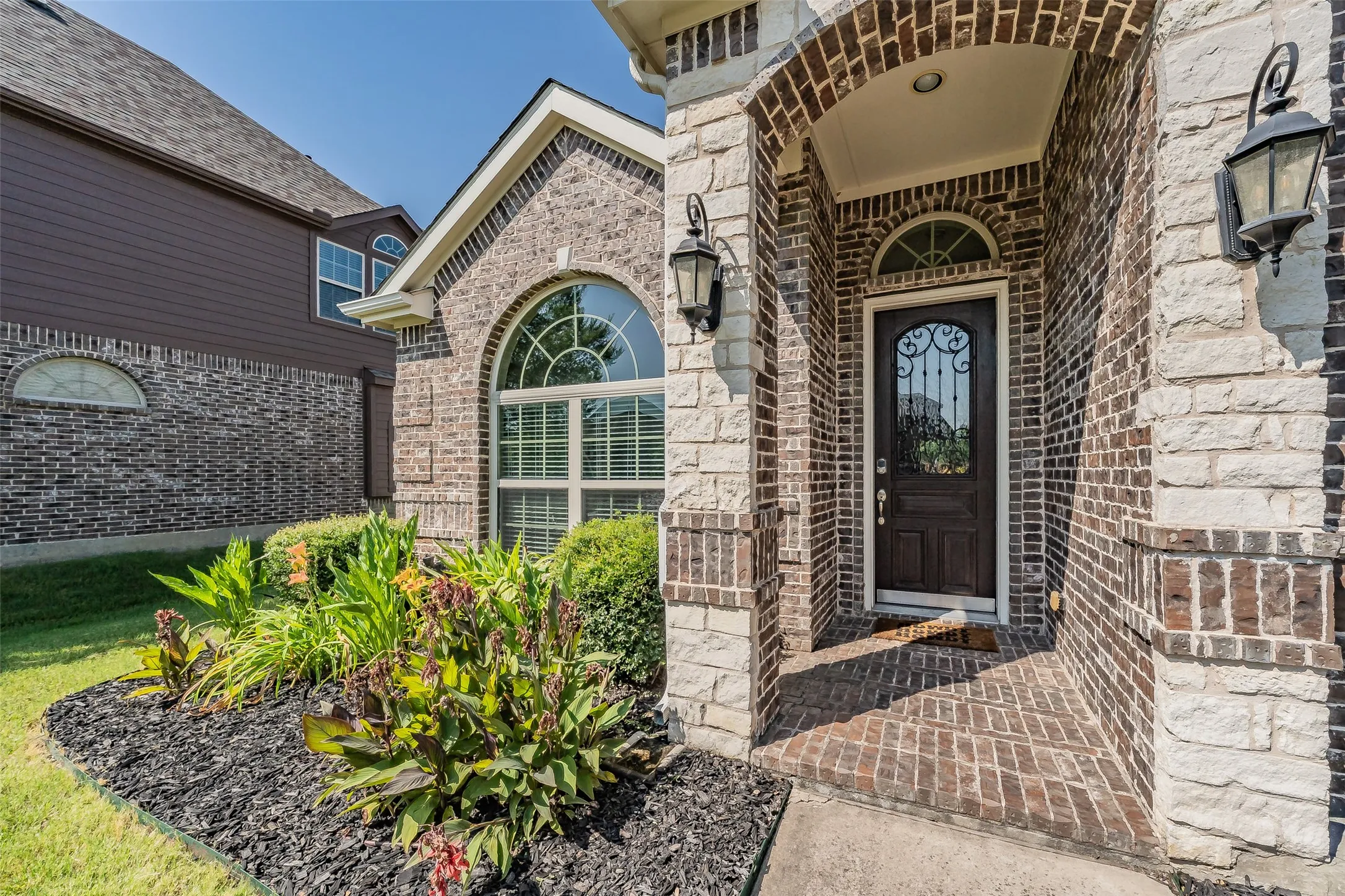 Brick and stone facade featuring an arched entryway, decorative glass front door, and two exterior lanterns