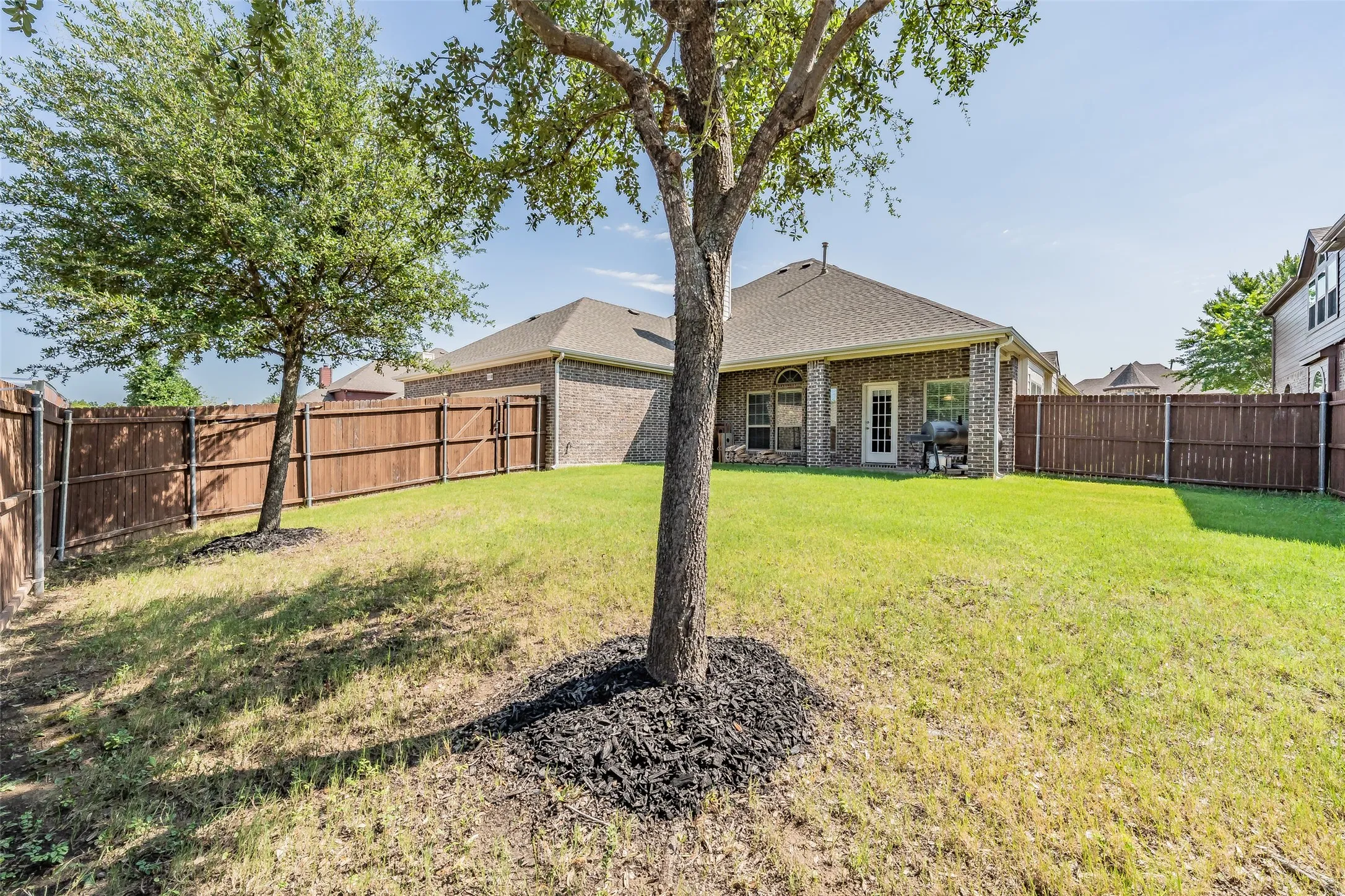 Spacious backyard featuring a privacy fence, mature trees, and a brick exterior with a covered patio