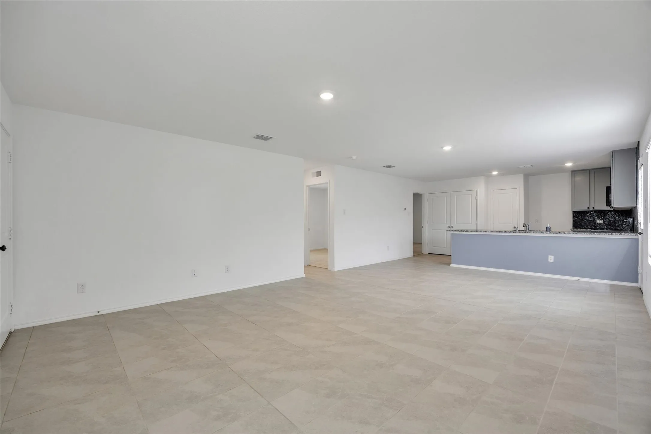 Spacious open-concept living area featuring light-toned tile flooring, recessed lighting, and a kitchen bar with a dark countertop