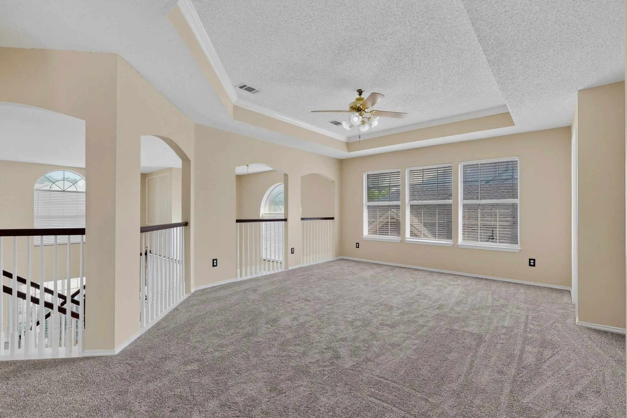Spacious upper-level room featuring plush carpeting, a tray ceiling with a brass-finish ceiling fan, and three large windows with blinds