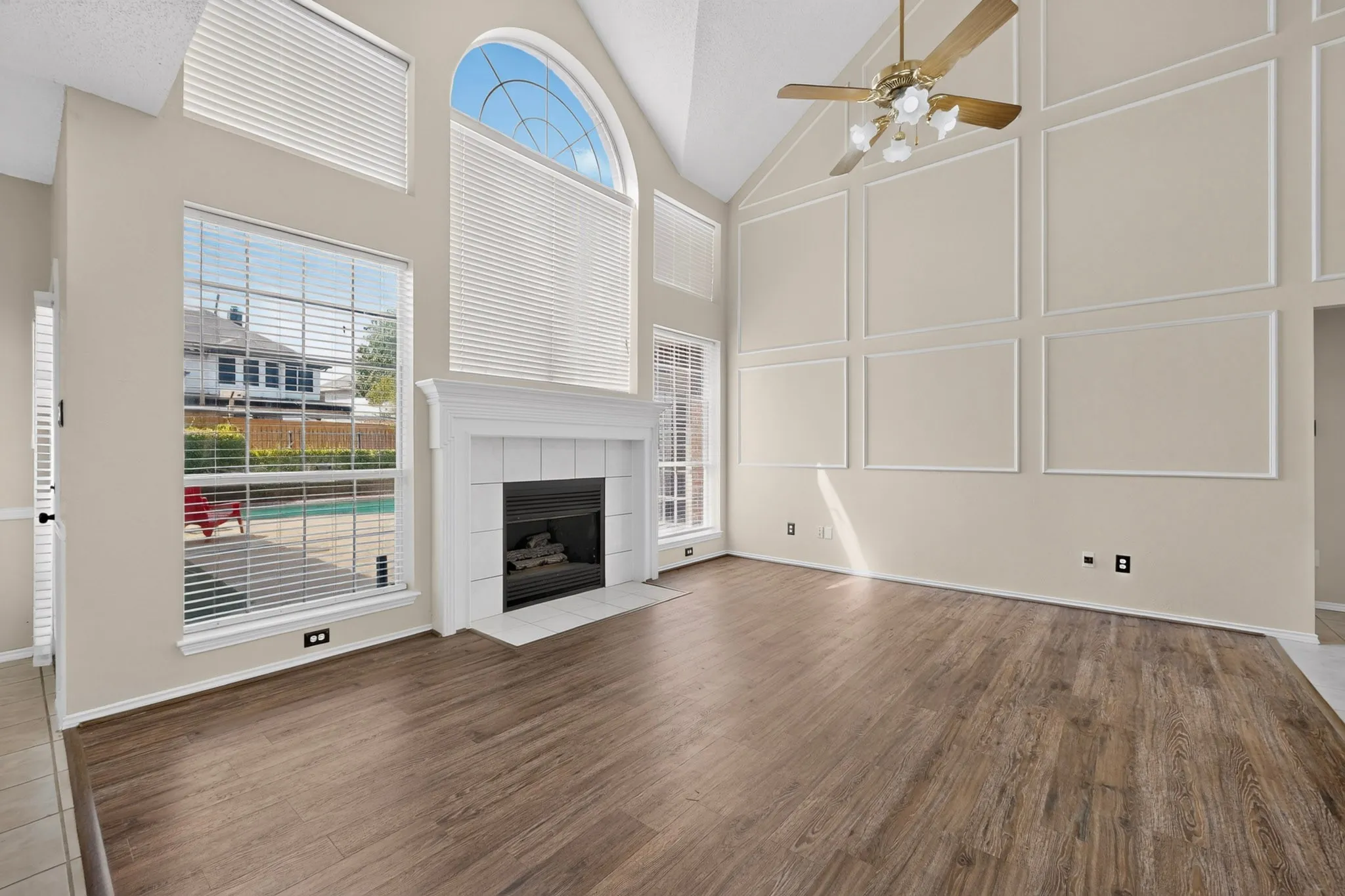Spacious living area featuring a vaulted ceiling with decorative trim, a fireplace with a white mantel, large windows, and durable wood-finish flooring