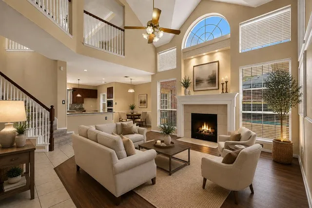 staged Soaring two-story living room featuring a gas fireplace with white mantel surround, hardwood flooring, and abundant natural light from multiple windows including a arched transom