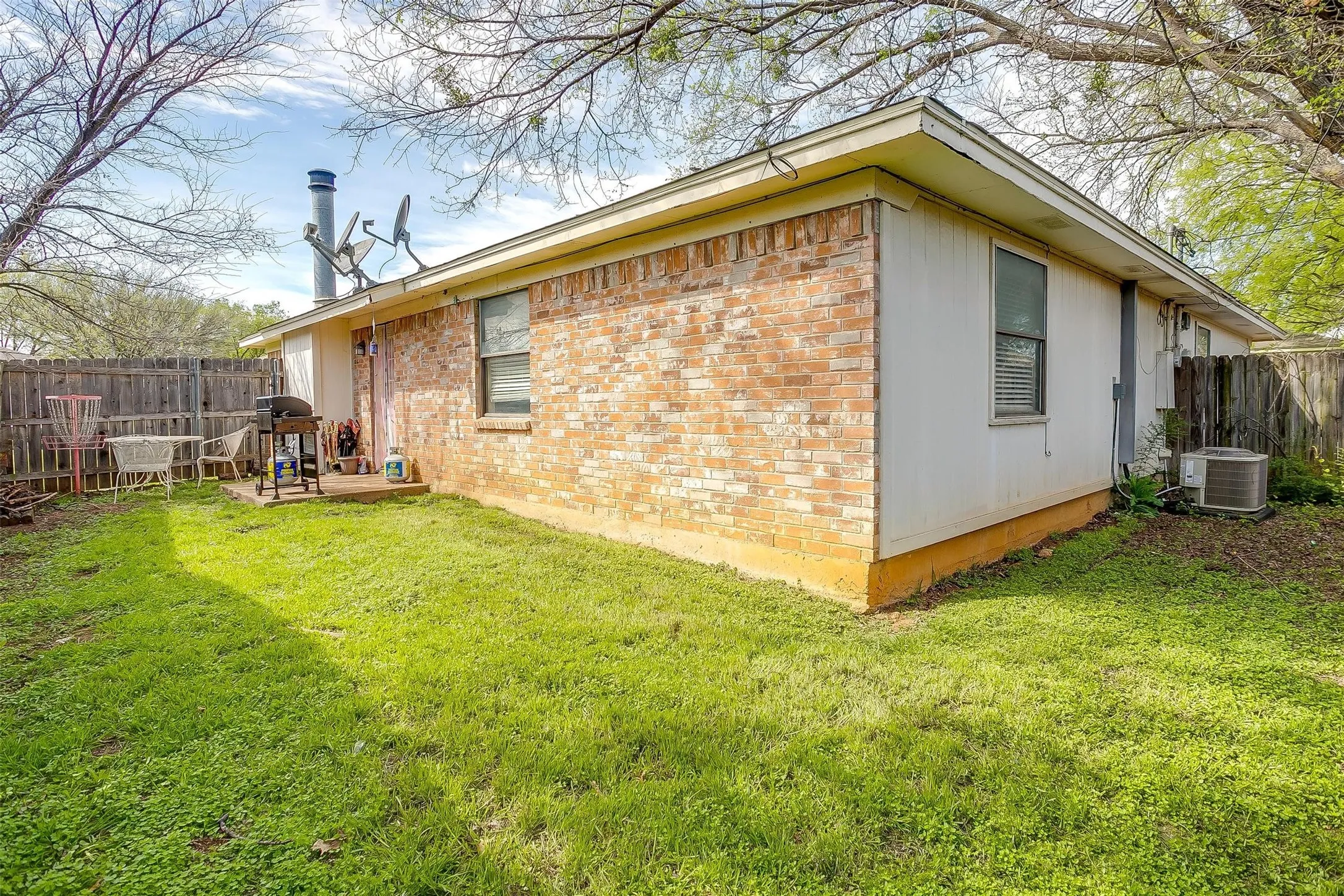 Exterior wall of house featuring a multi-colored brick in shades of red, orange, tan and cream, and backyard featuring established grass and concrete pad.