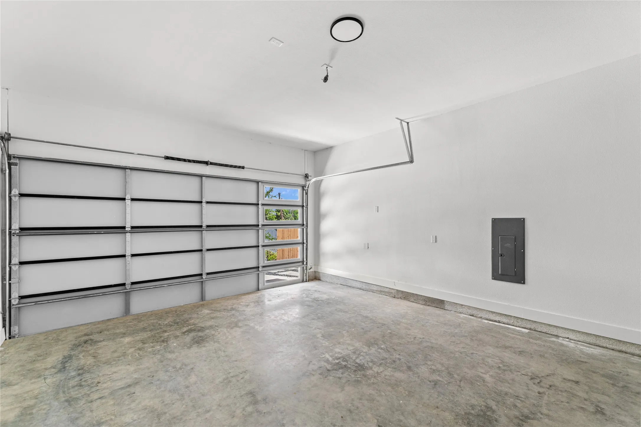Garage space featuring a frosted panel garage door with clear window accents, concrete flooring, crisp white walls, a visible electrical panel, and a ceiling-mounted light fixture