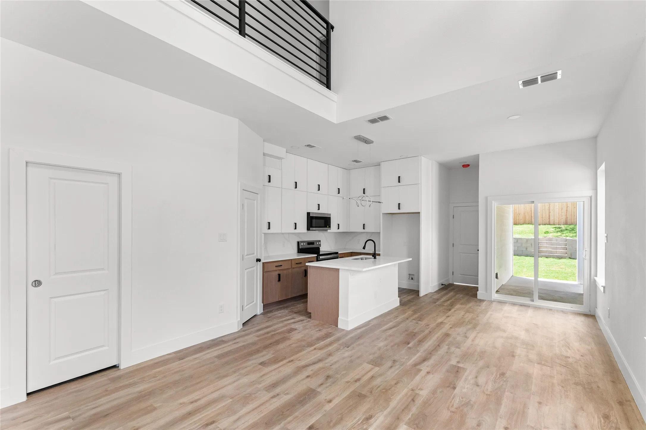 Open-concept living area featuring high ceilings, wood-finish flooring, a contemporary kitchen with a central island and white cabinetry, and sliding glass doors leading to the outdoor space