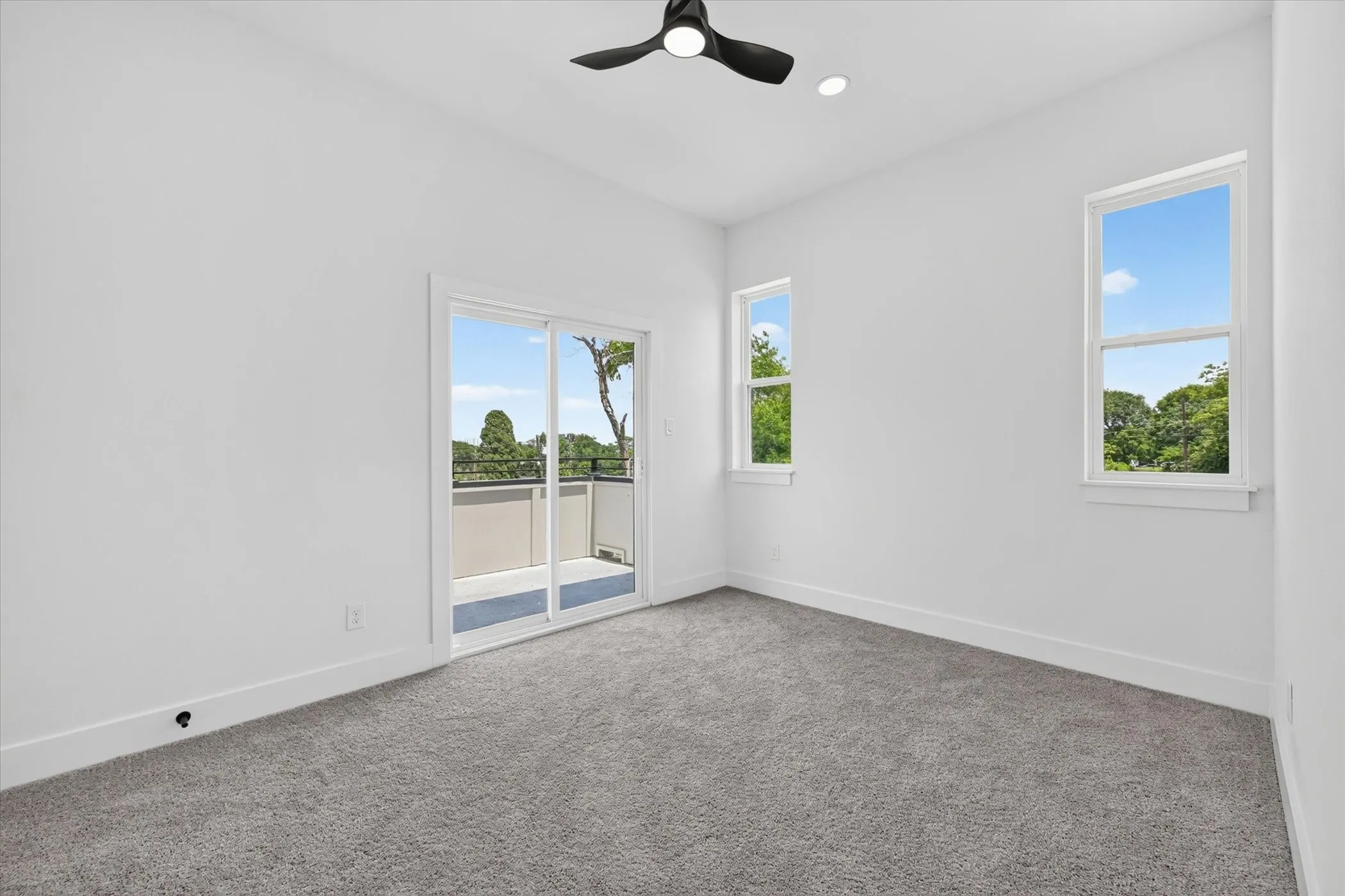 Spacious room featuring light gray carpeting, crisp white walls, a modern black ceiling fan, recessed lighting, and a sliding glass door leading to an outdoor balcony