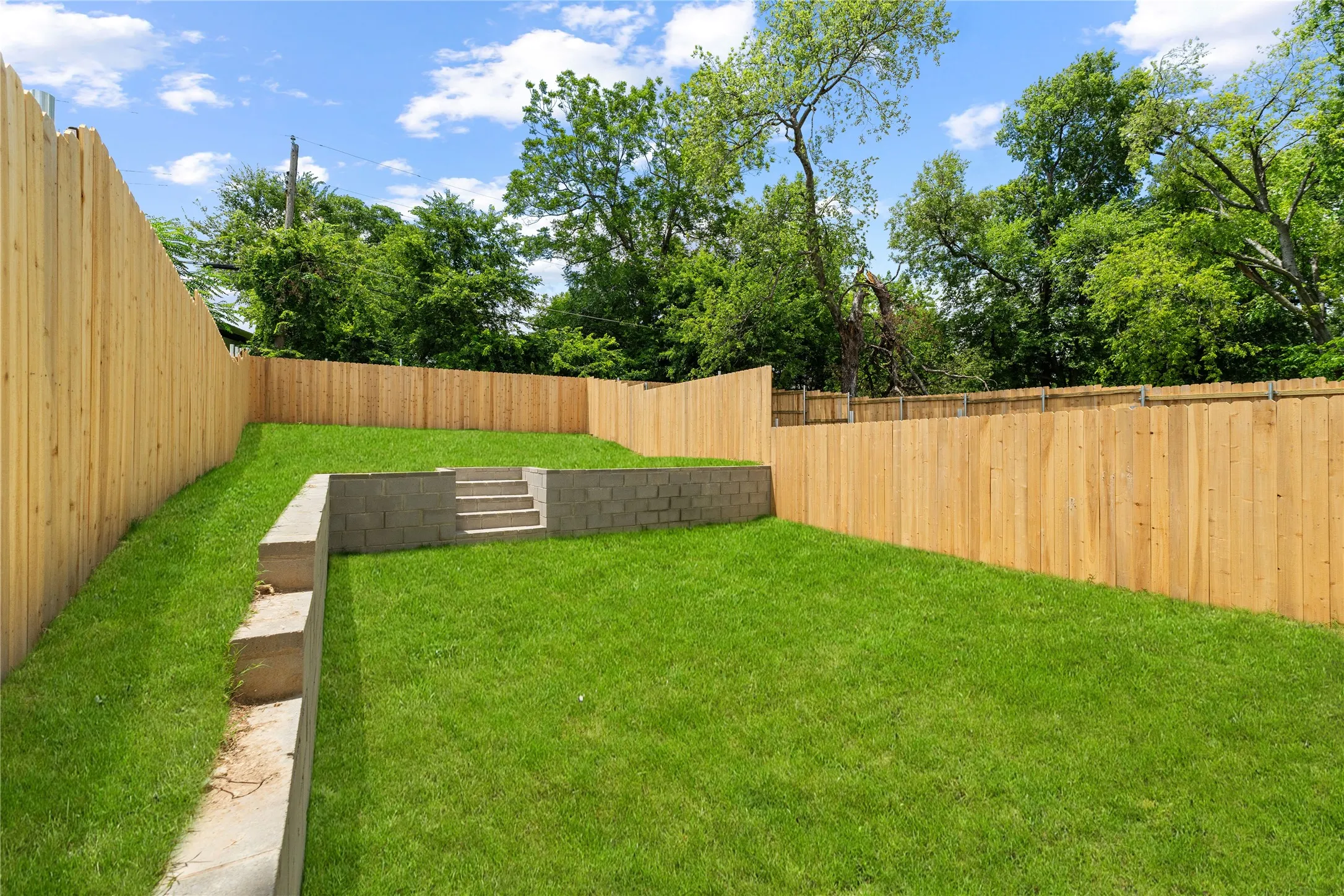 Expansive backyard featuring a manicured lawn, a new wood privacy fence, concrete block retaining walls, and integrated concrete block steps