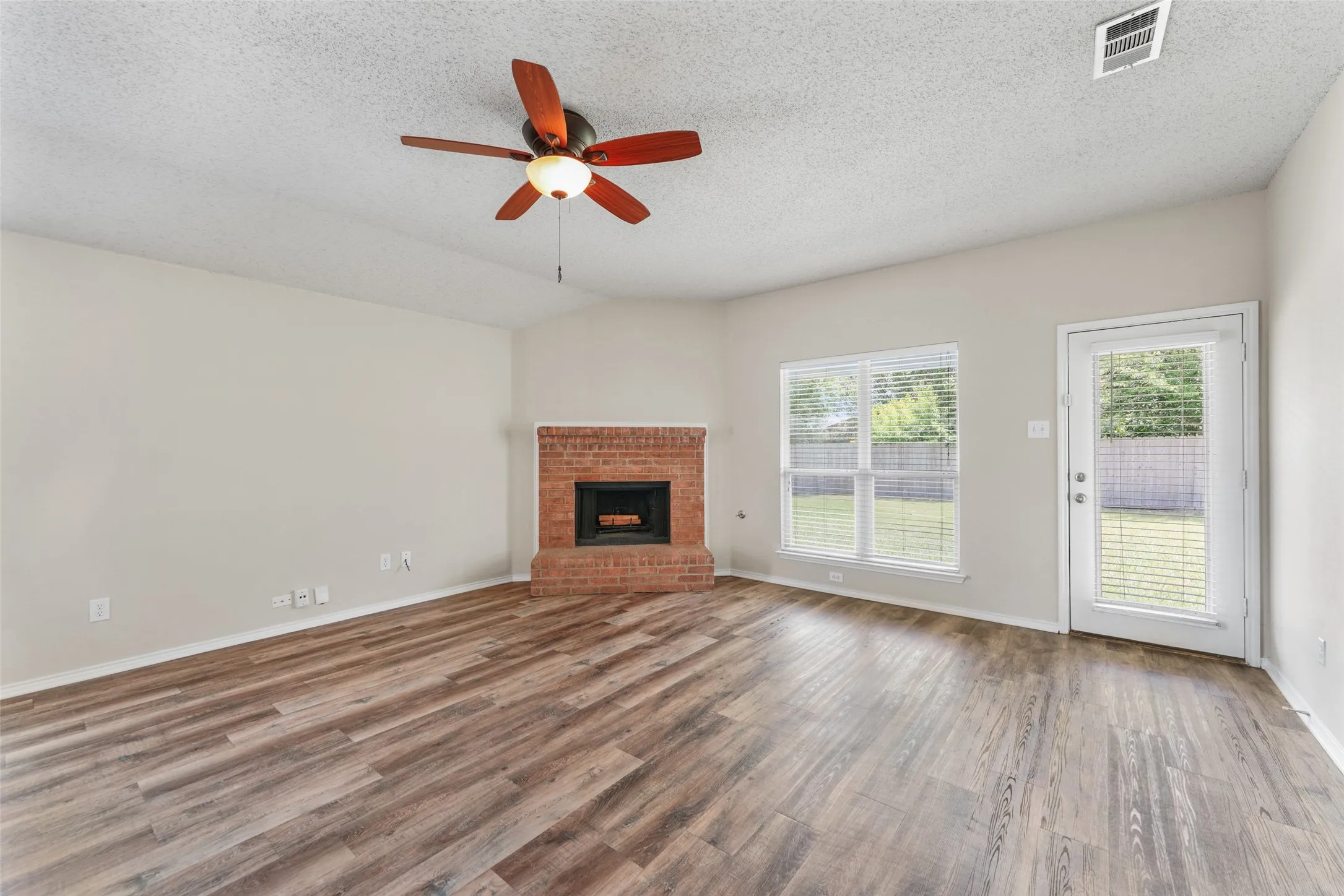 Living room featuring wood-finish flooring, a brick fireplace, a ceiling fan with light fixture, and a glass-paned door opening to the exterior