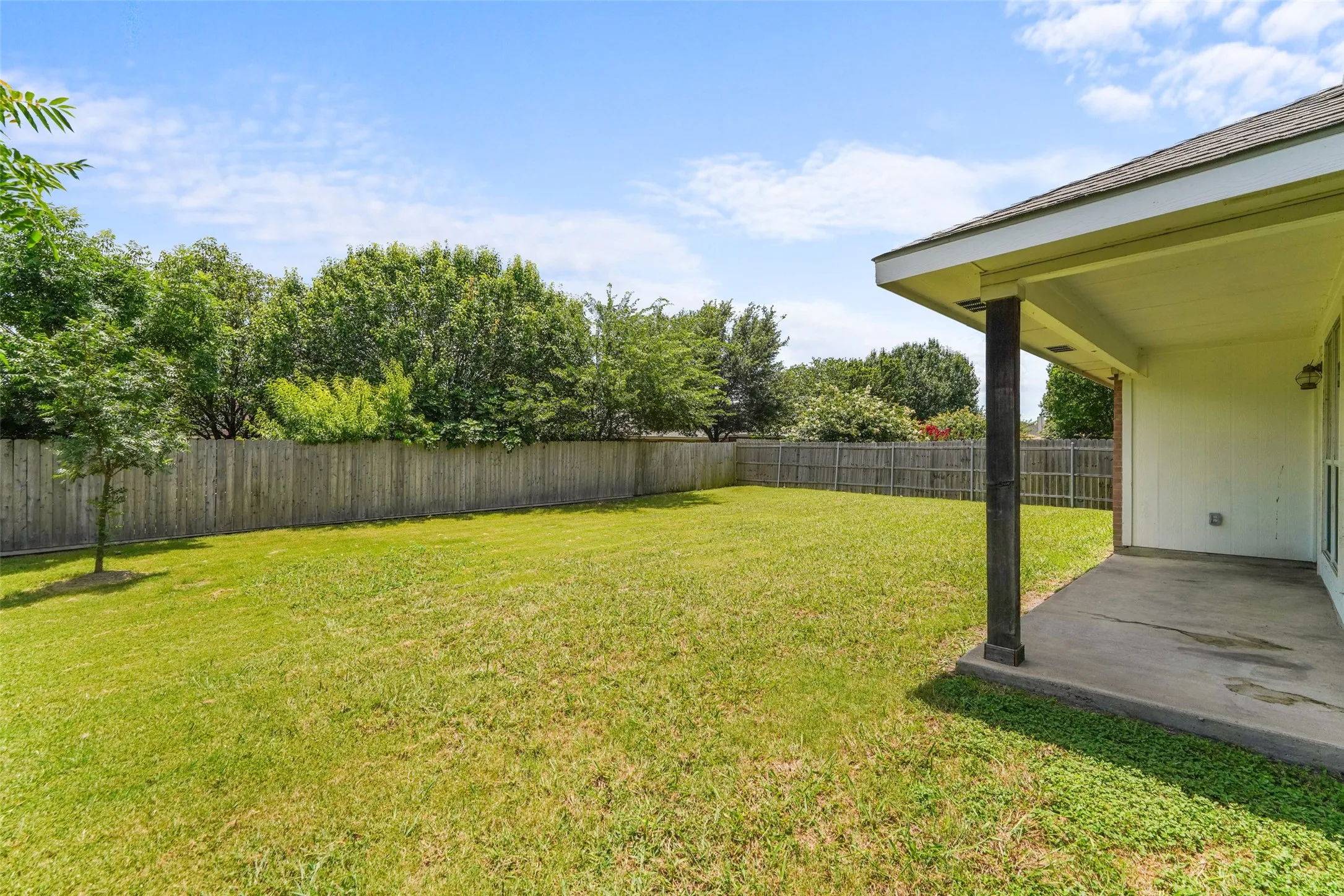 Expansive backyard featuring a large grass lawn, perimeter wood fencing, mature trees, and a covered patio with a concrete slab