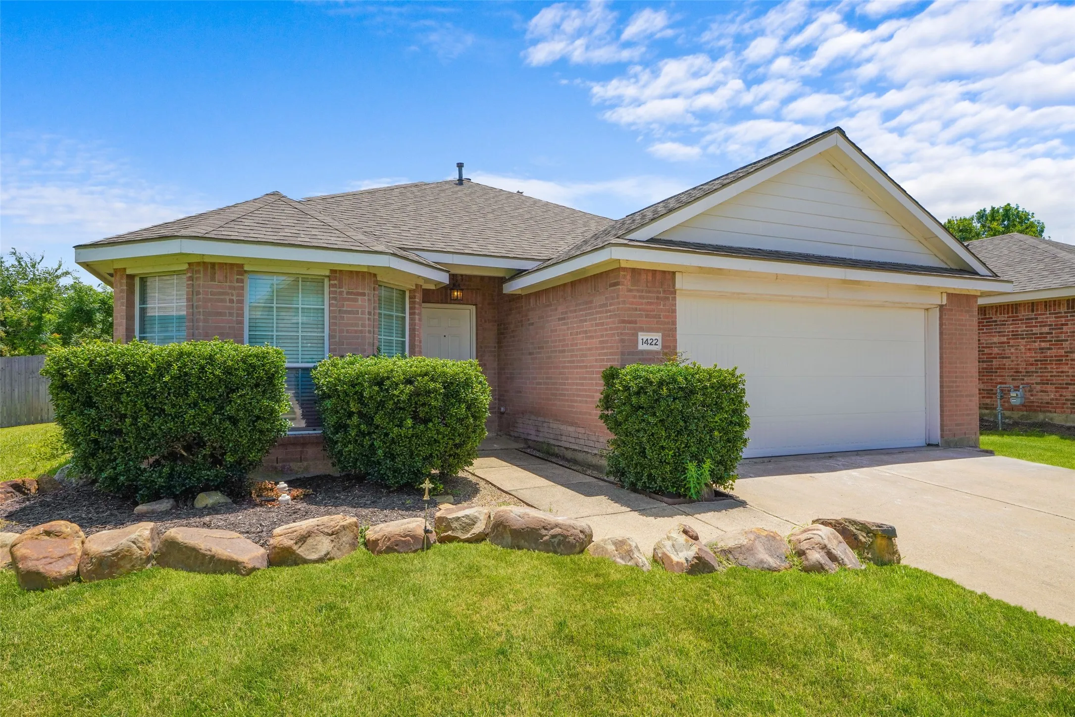 Brick exterior with a peaked roofline, an attached two-car garage, bay windows, a concrete driveway, and a front yard featuring a rock border and mature shrubbery