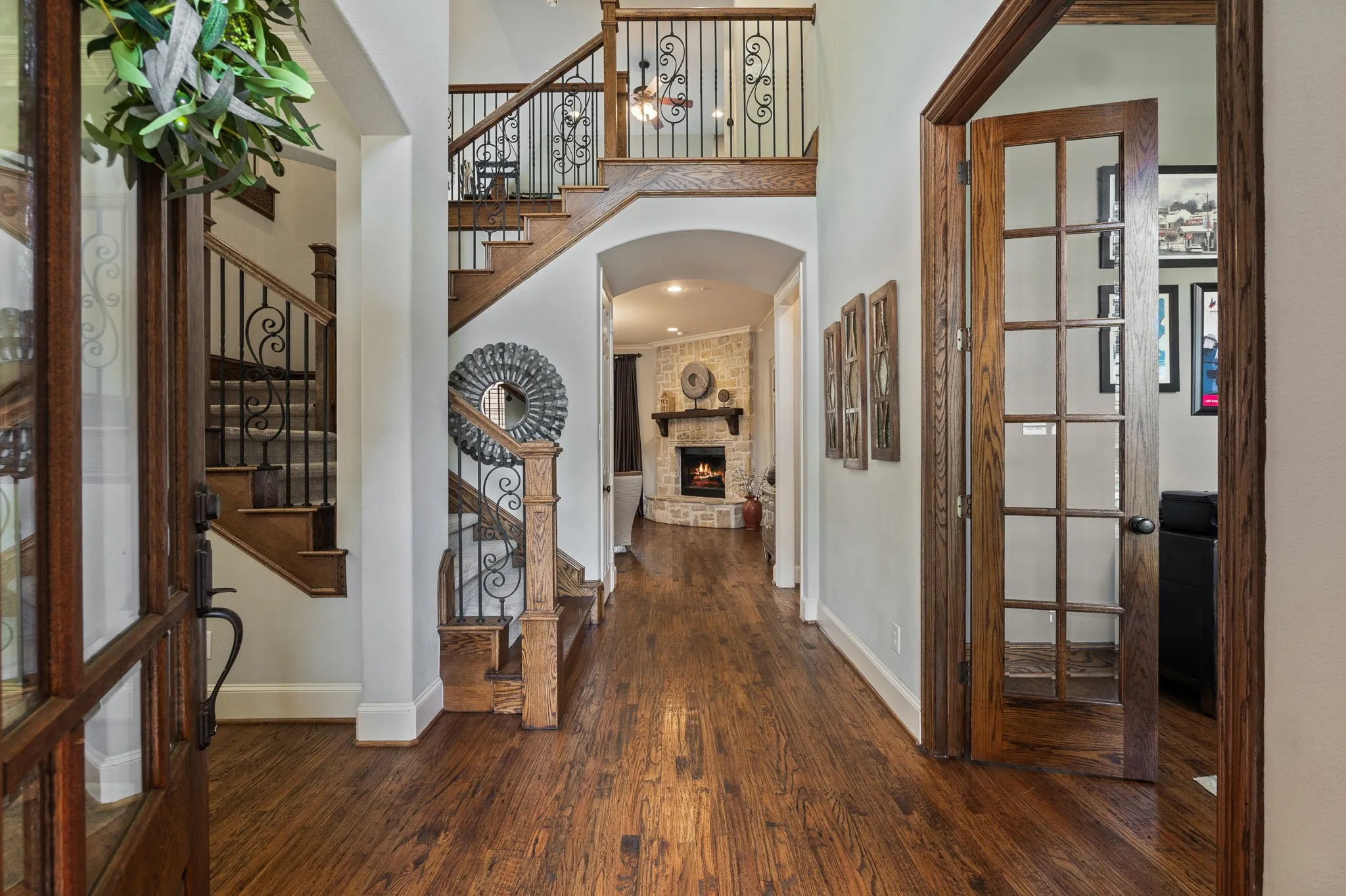 Grand entry foyer featuring rich wood-finish flooring, a prominent staircase with wrought iron balusters, a second-story balcony overlook, and a paneled glass door leading to an additional room