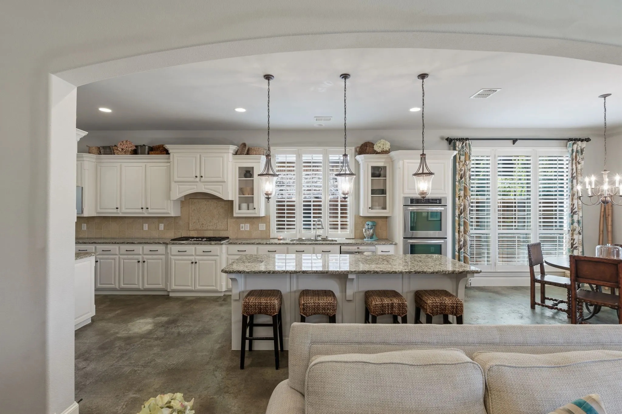 White cabinetry, integrated double ovens, an island with countertop seating, recessed lighting, and large windows with plantation shutters