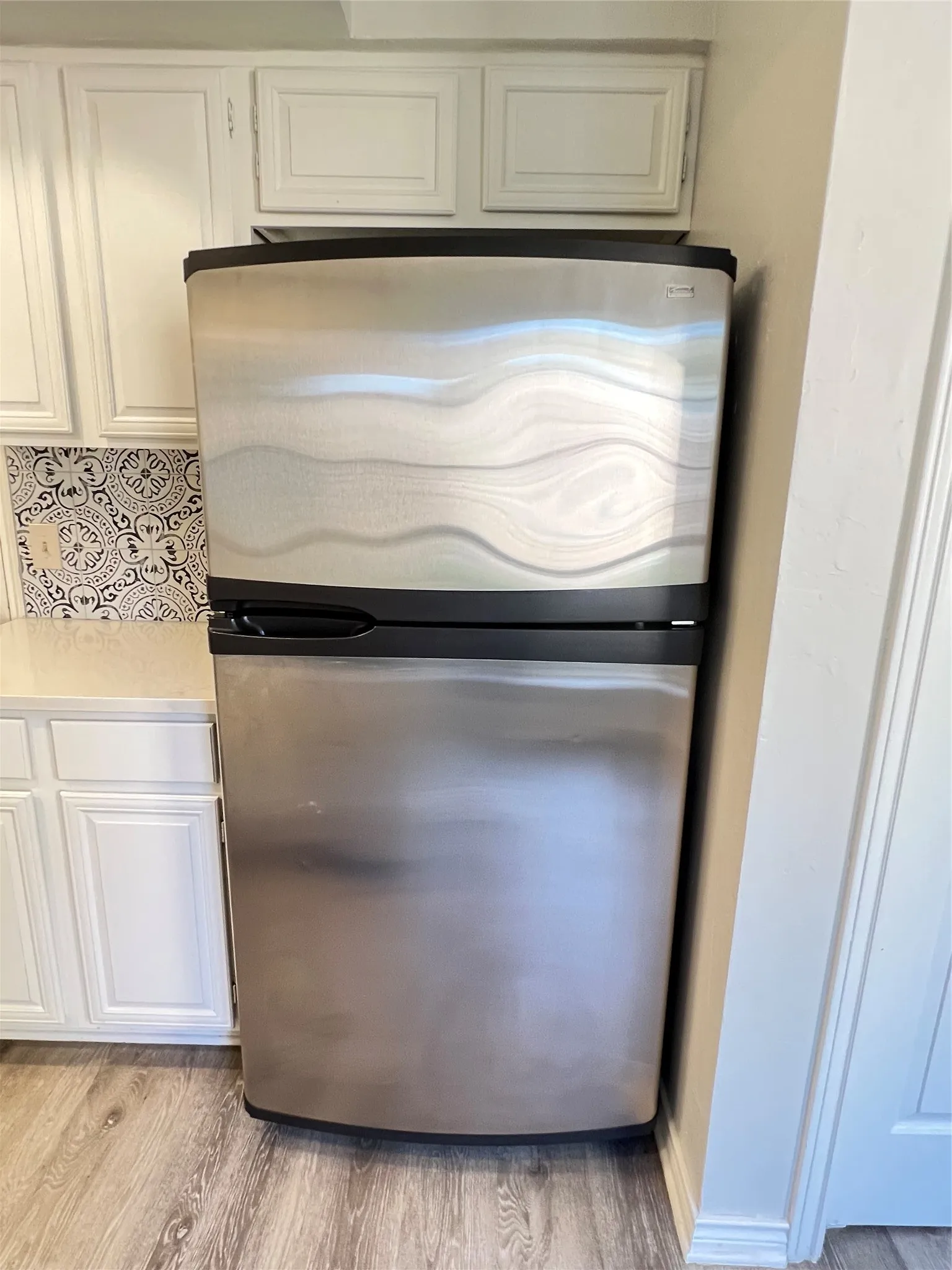 Stainless steel refrigerator appliance, white upper and lower cabinetry with paneling, patterned tile backsplash, light-toned countertop, and wood-finish flooring