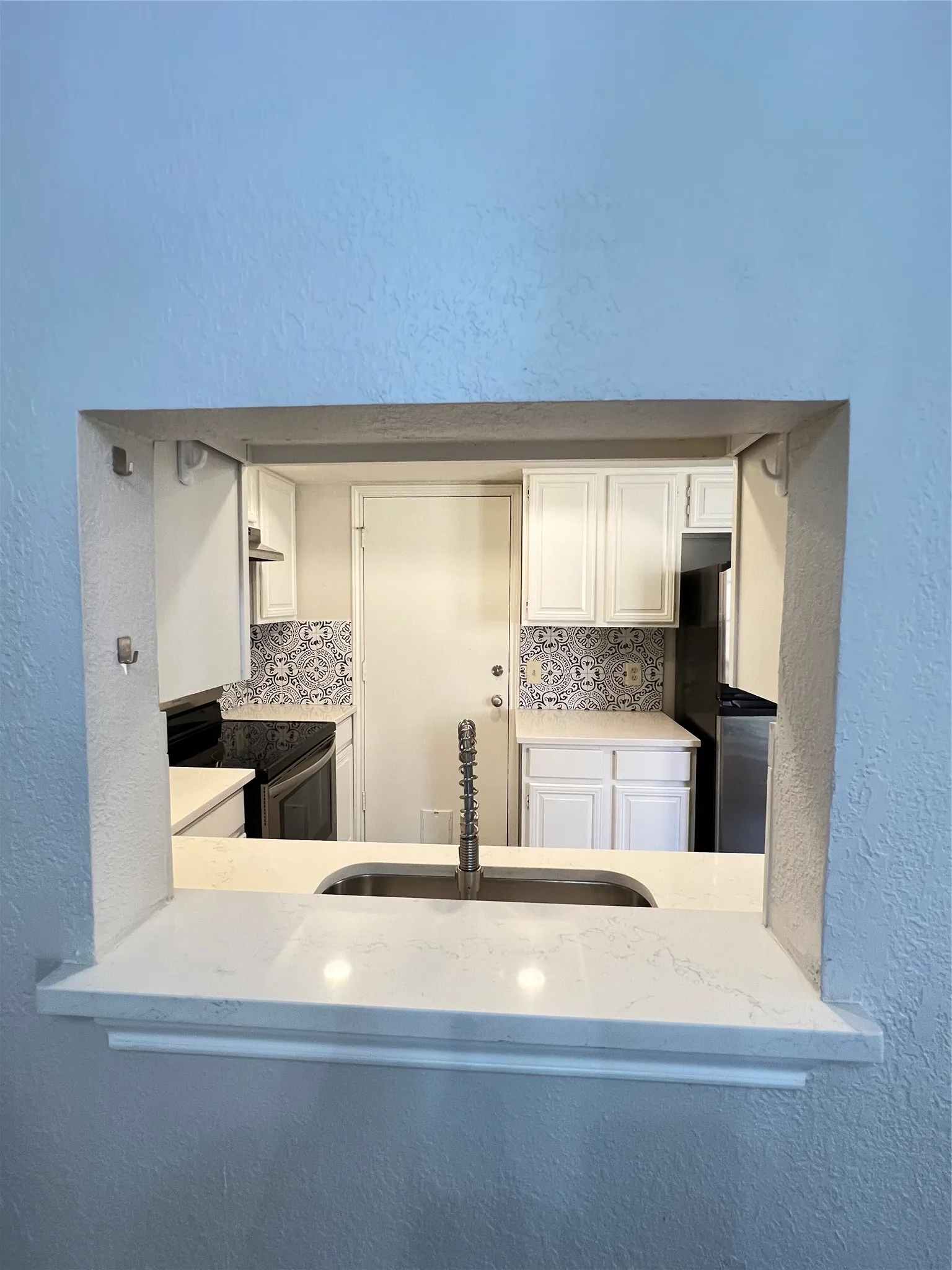 Kitchen pass-through featuring a stainless steel sink with a high-arc faucet, quartz countertops, white cabinetry, and a decorative tile backsplash