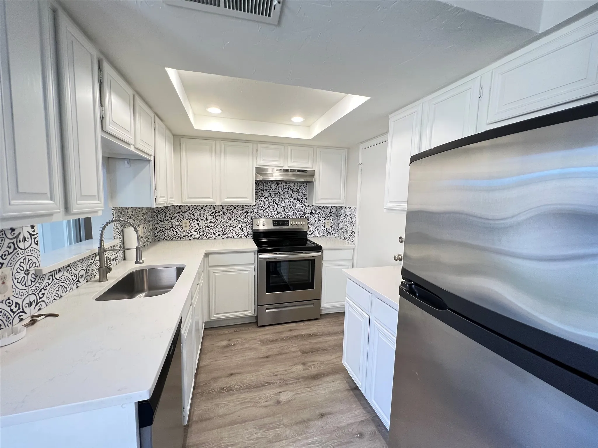 Kitchen featuring white shaker cabinetry, patterned tile backsplash, stainless steel appliances, light-toned countertops, and wood-finish flooring