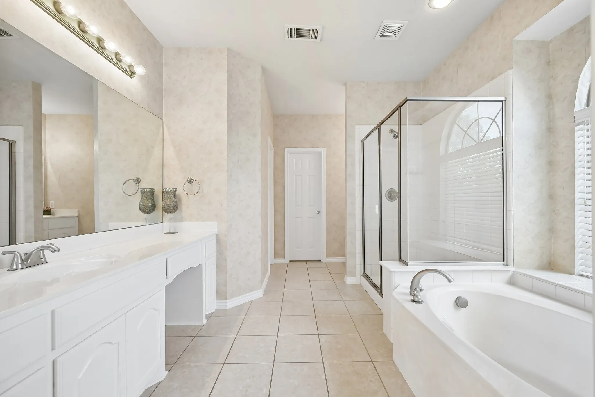 Spacious bathroom featuring a dual vanity with integrated sink, a large soaking tub, a separate glass-enclosed shower, arched window with privacy blinds, and tile flooring