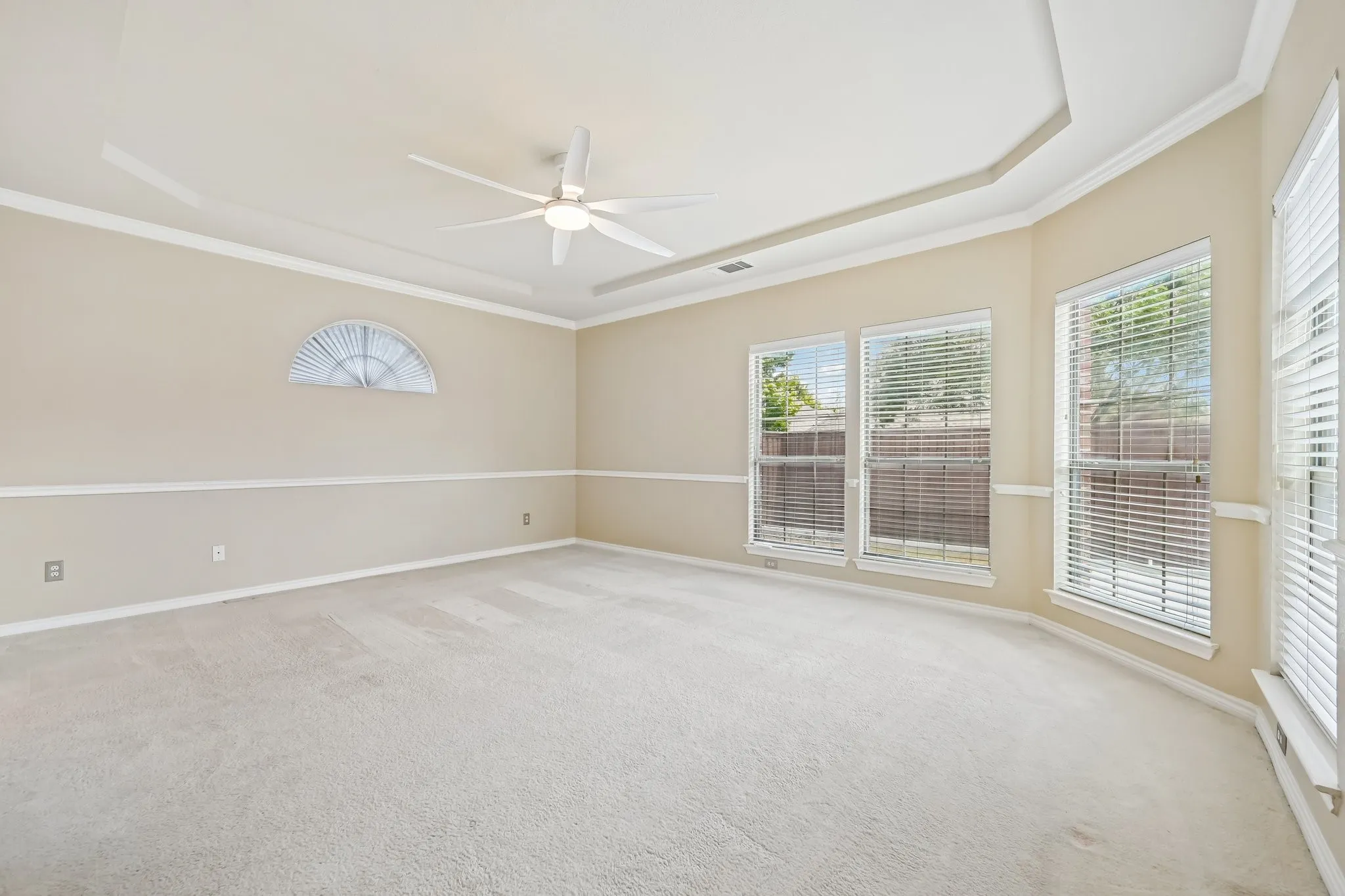 Spacious room featuring light-colored carpet, a tray ceiling with a modern ceiling fan, multiple windows with horizontal blinds, and a half-round window