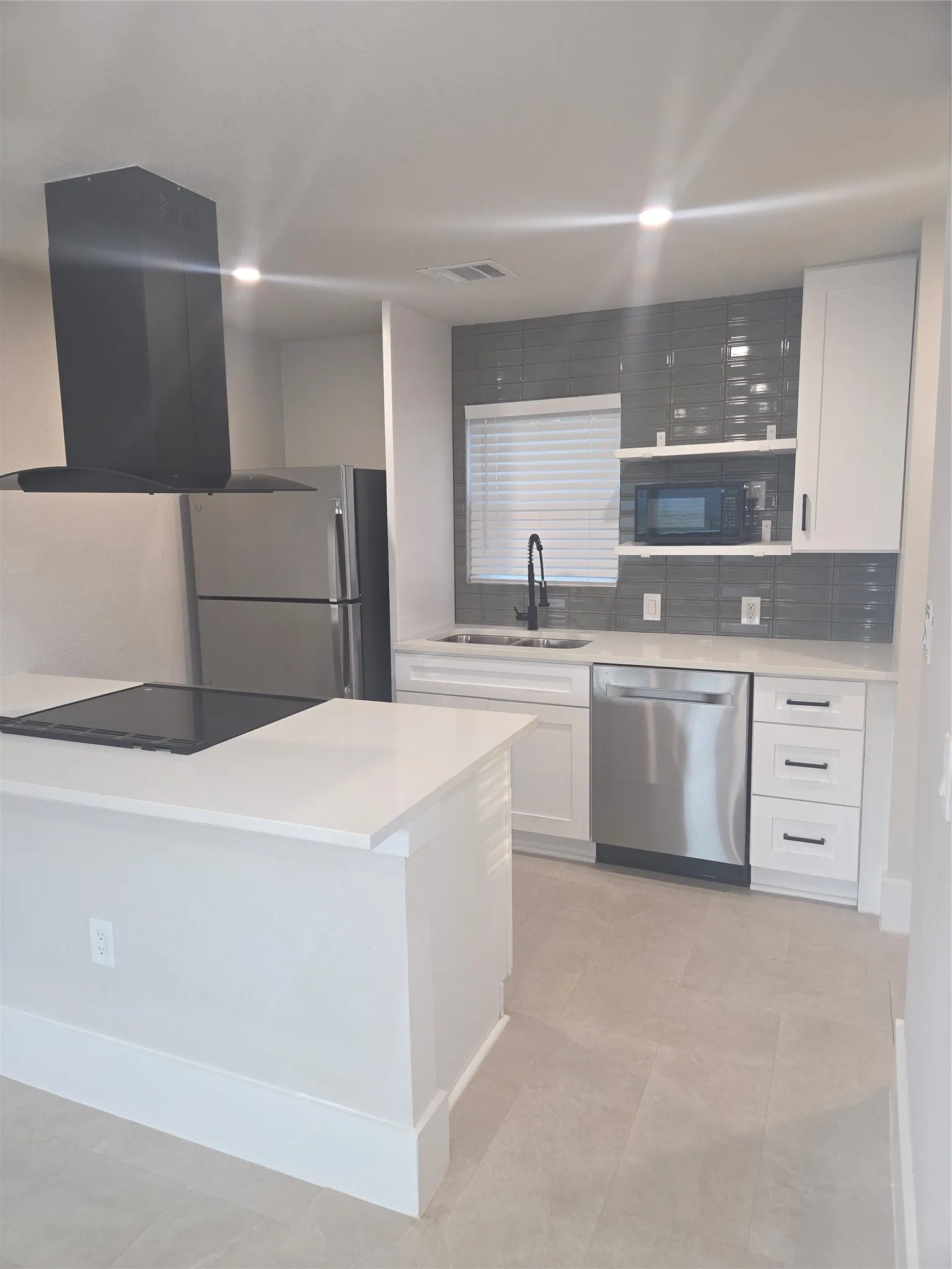Modern kitchen featuring an island with an integrated cooktop, a black range hood, stainless steel appliances, white cabinetry, and gray subway tile backsplash