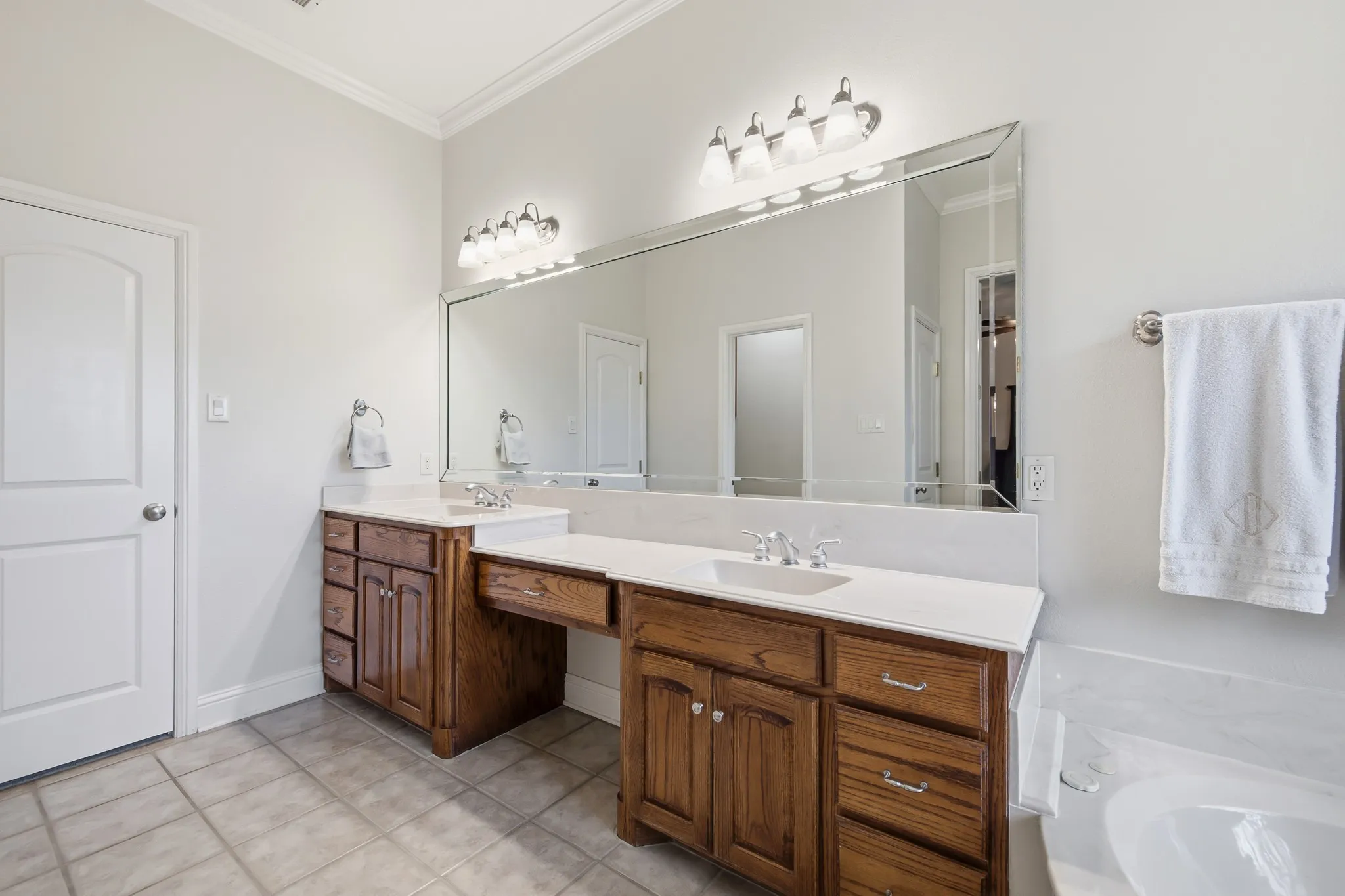 Bathroom featuring dual wood-finish vanities with undermount sinks and light-toned countertops, a large beveled mirror, brushed nickel fixtures, and tile flooring