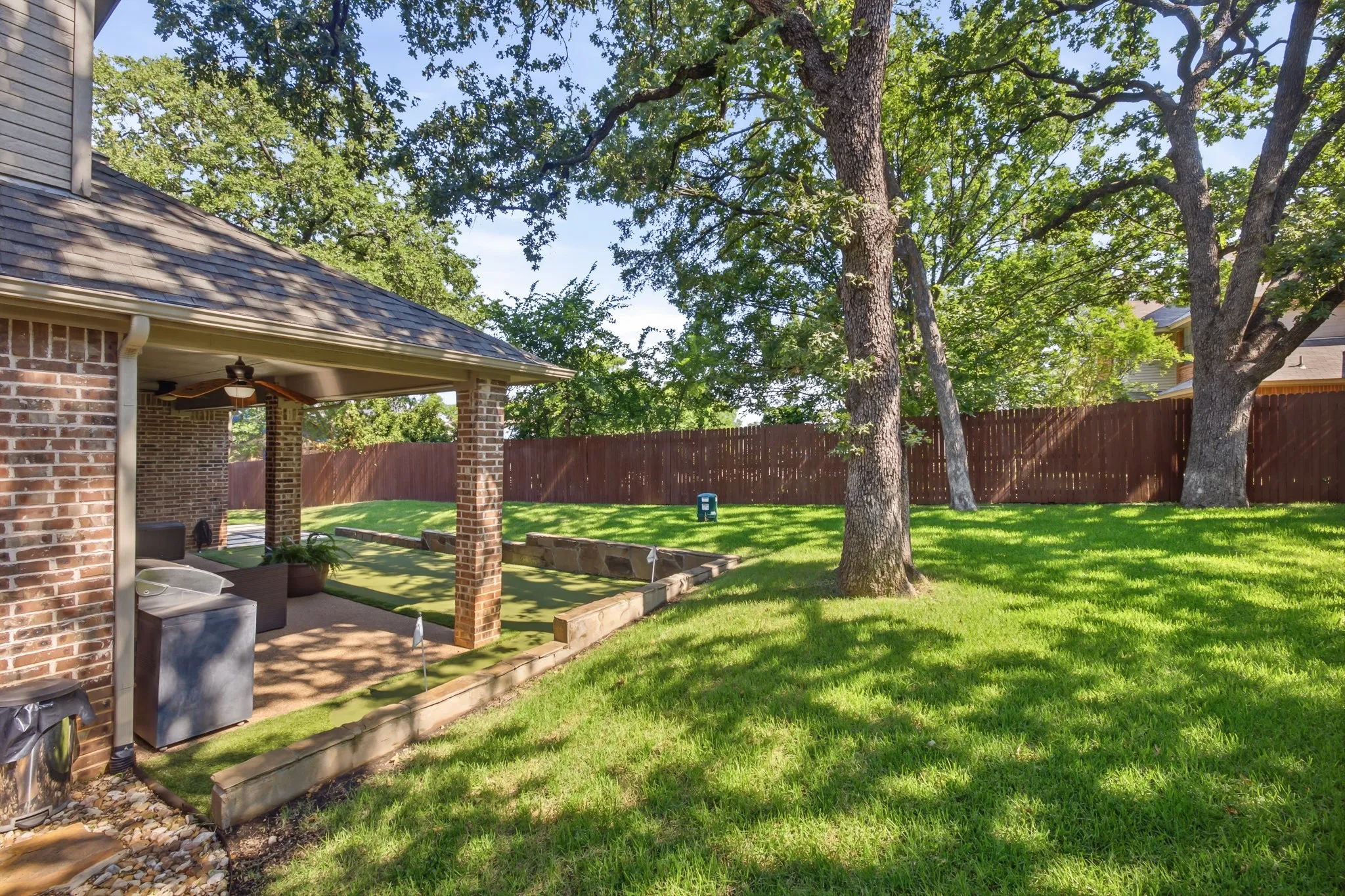 Beautiful backyard featuring a covered patio with brick columns and a ceiling fan, mature trees, a privacy fence, and tiered landscaping beds