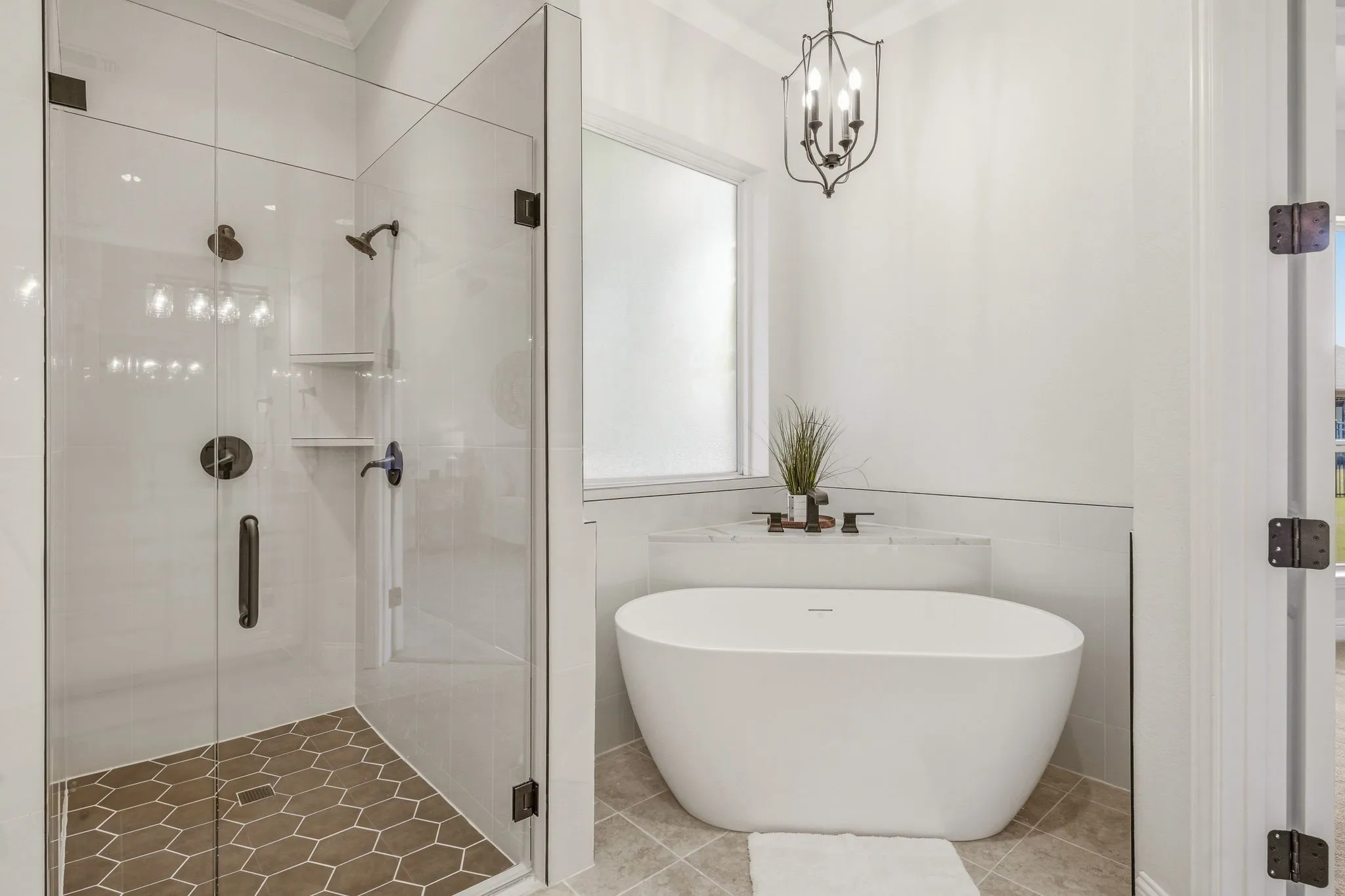 Modern primary bathroom featuring a freestanding soaking tub with a deck-mounted faucet, a glass-enclosed shower with hexagonal floor tiles, a window with privacy glass, and a black metal chandelier