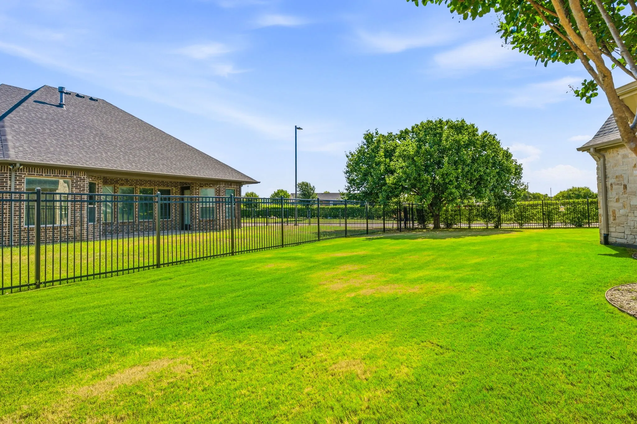 Expansive grassy yard enclosed by a black metal fence, featuring a mature shade tree and a brick exterior building with multiple windows