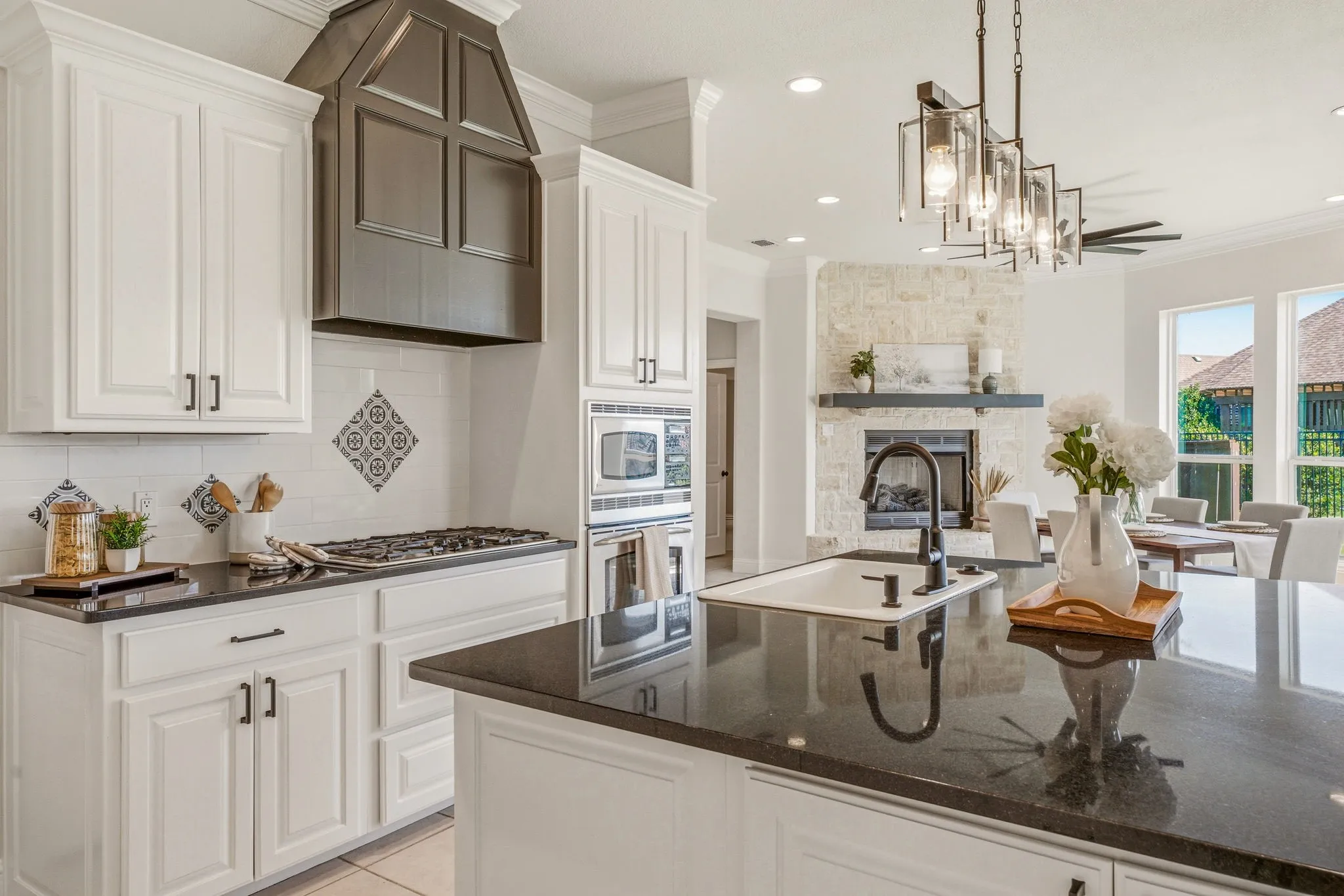 Open-concept kitchen featuring white cabinetry, dark countertops, built-in stainless steel appliances, a center island with sink, and a contrasting dark range hood