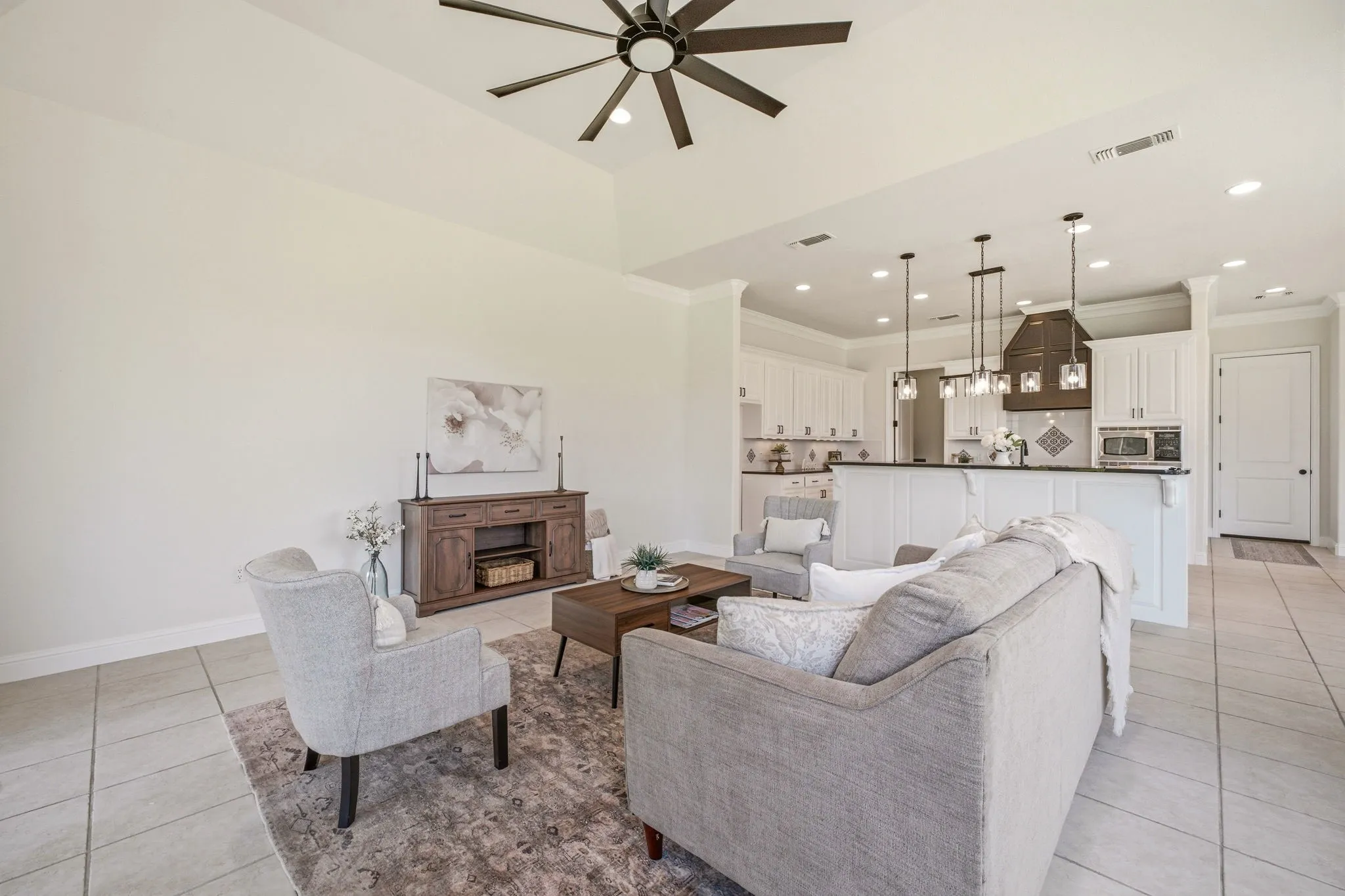 Vaulted ceiling living area featuring a dark wood-finish ceiling fan, neutral tile flooring, and an open-concept kitchen with white cabinetry, stainless steel appliances, and pendant lighting