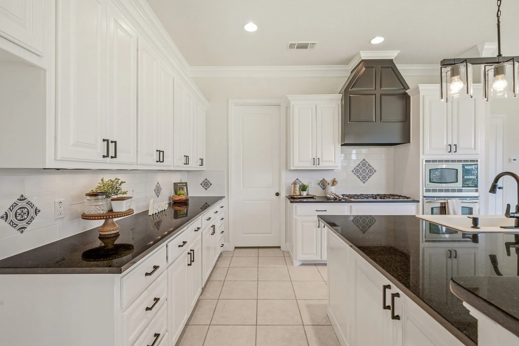 Bright kitchen featuring extensive white cabinetry, dark countertops, a contrasting dark wood range hood, a decorative tile backsplash, and a central island with a sink