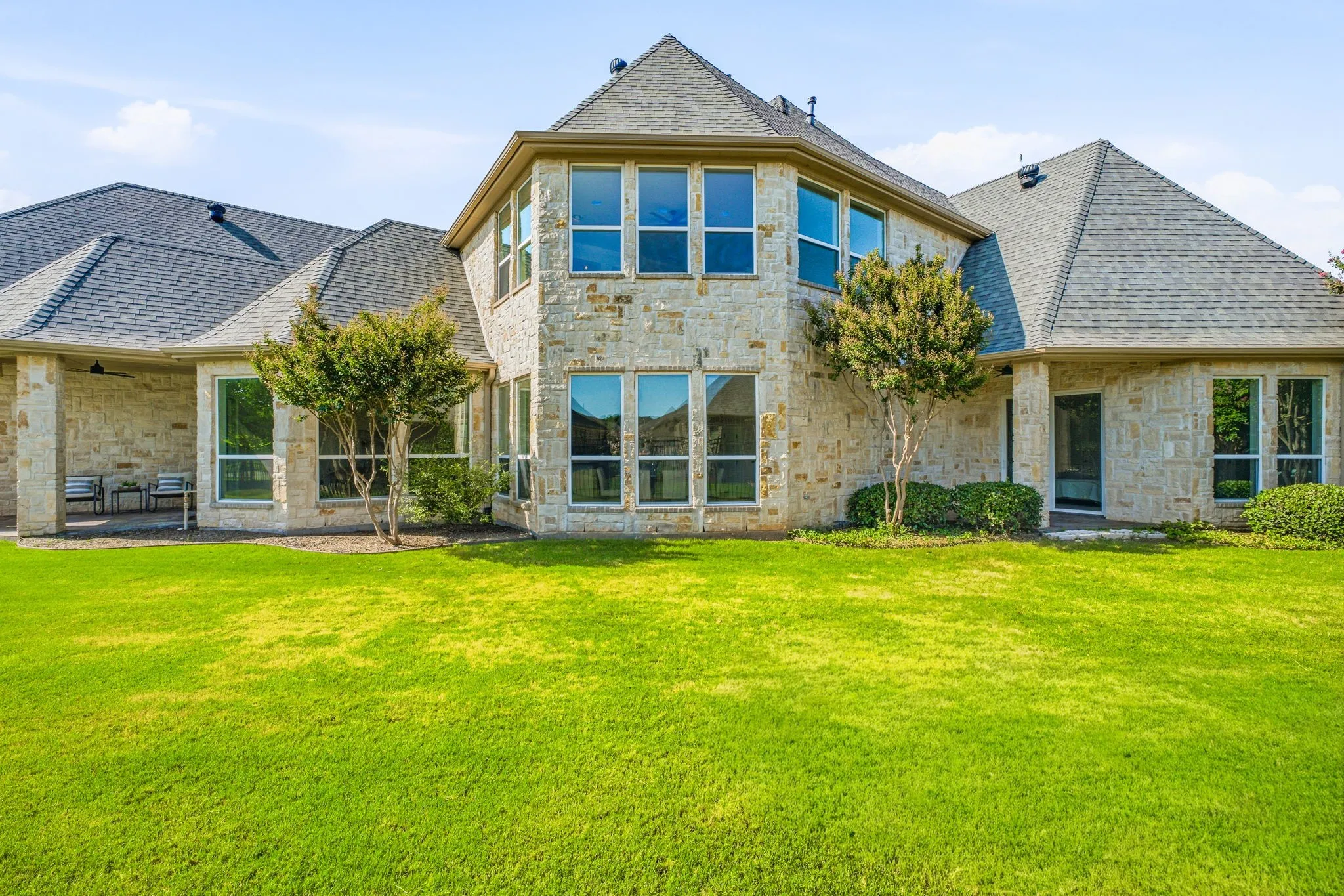 Expansive rear exterior featuring a stone facade, multiple large windows, a covered patio area, and a lush green lawn