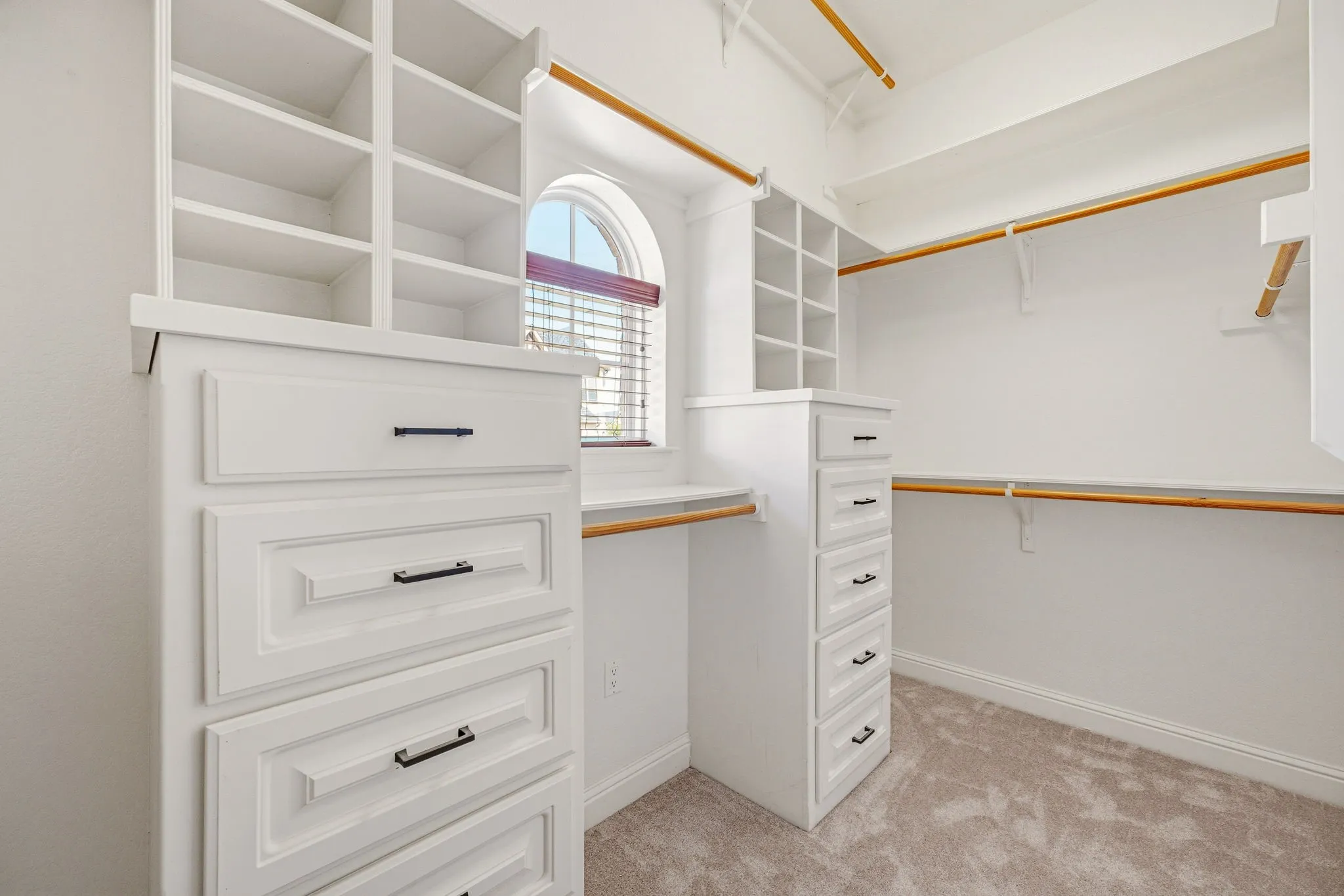 Primary Custom built-in white cabinetry with drawers and shelving, complemented by an arched window with blinds, wood-finish hanging rods, and light-toned carpeting