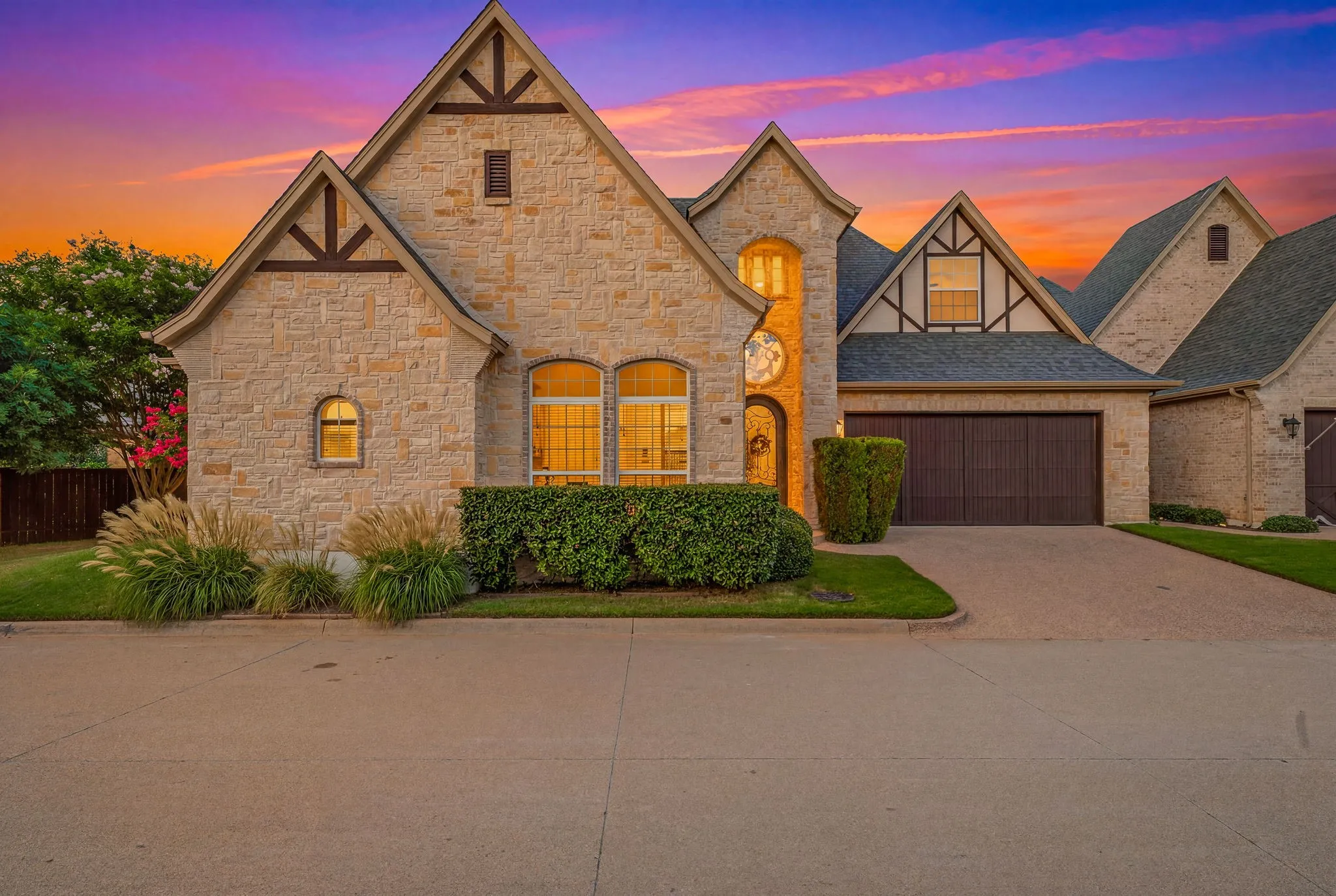 Stone exterior residence featuring pitched gables, dark trim accents, a two-car garage, manicured shrubbery, and a concrete driveway