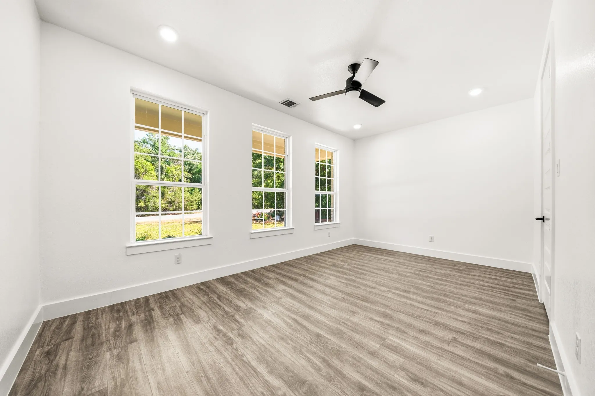 Spacious room featuring three double-hung windows, wood-finish flooring, recessed lighting, a ceiling fan, and white baseboards