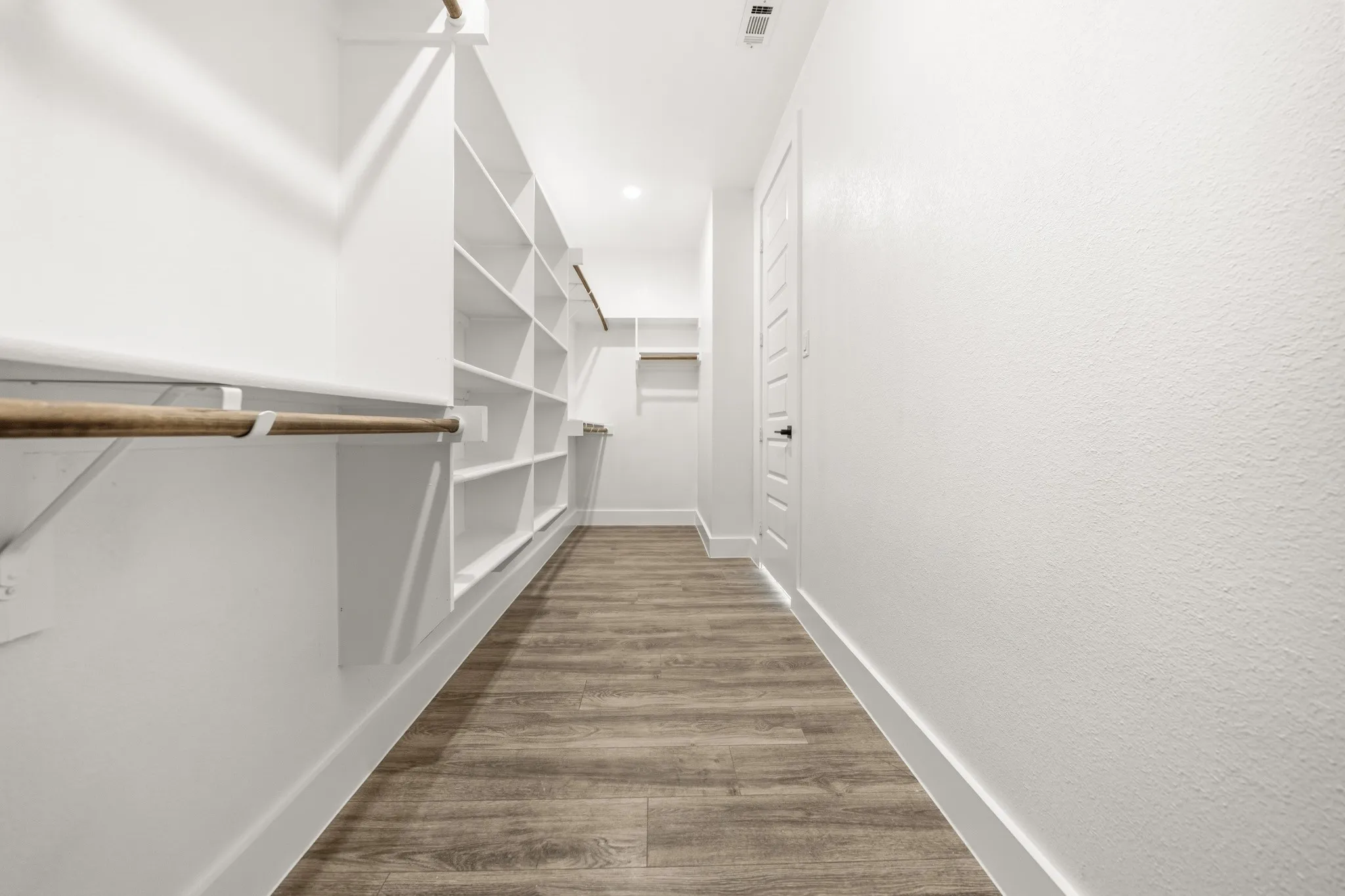 Elongated closet featuring wood-finish flooring, white built-in shelving, white walls, and a recessed ceiling light