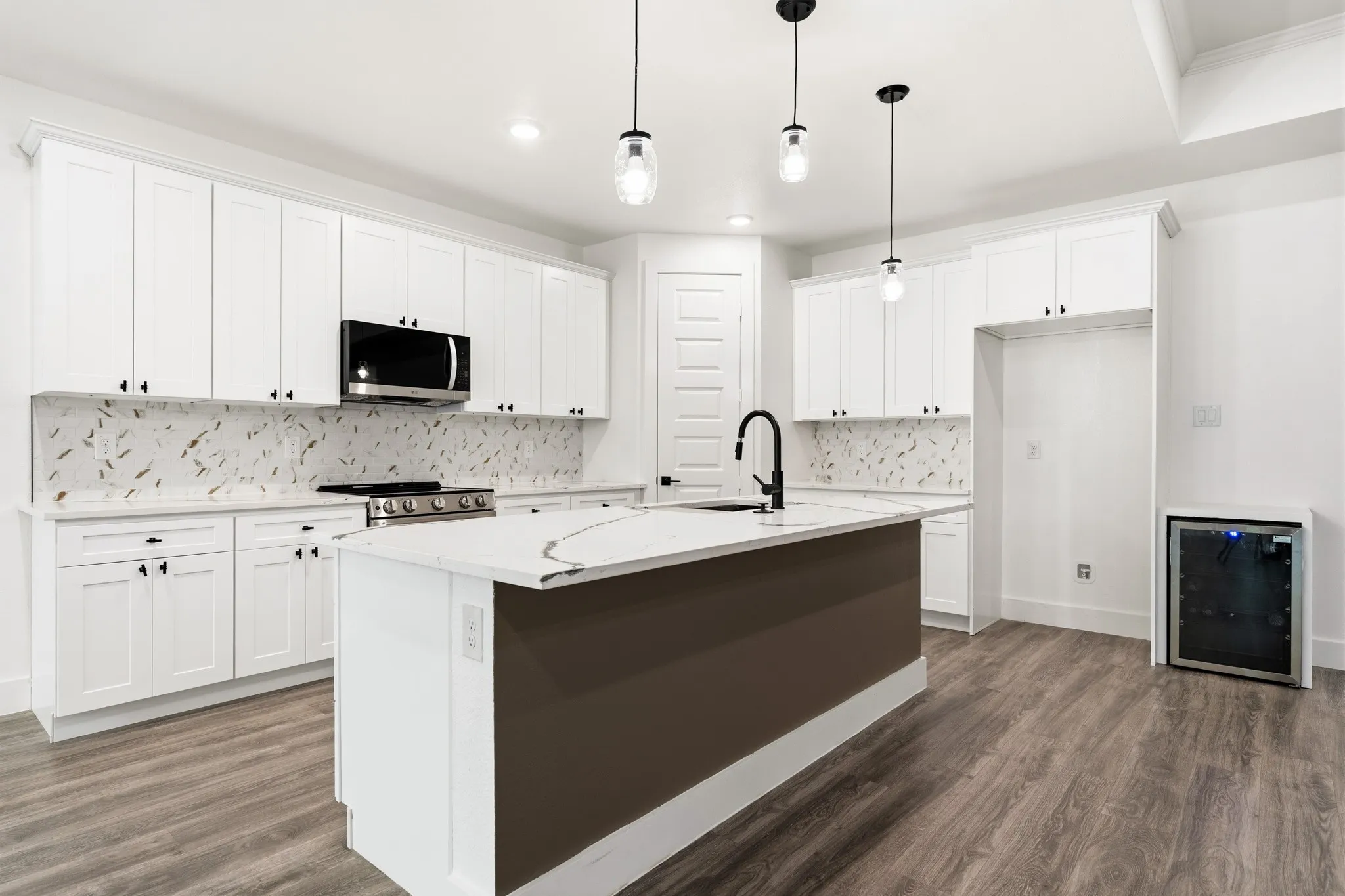 Open-concept kitchen featuring an island with a built-in sink, white cabinetry with black hardware, patterned tile backsplash, and wood-finish flooring