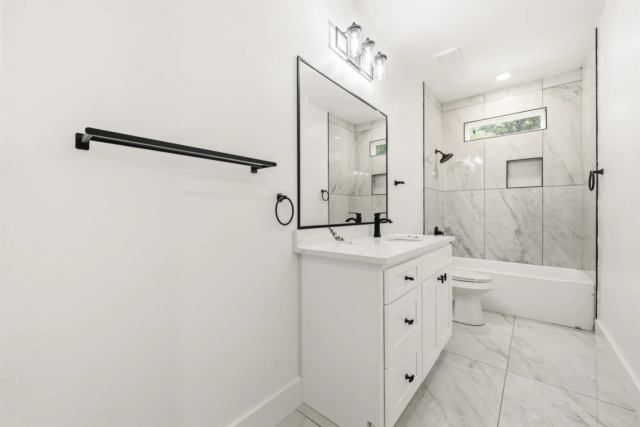 Bathroom featuring a white vanity with black hardware, a rectangular mirror, a bathtub with a tiled surround, a recessed shower niche, and a horizontal window