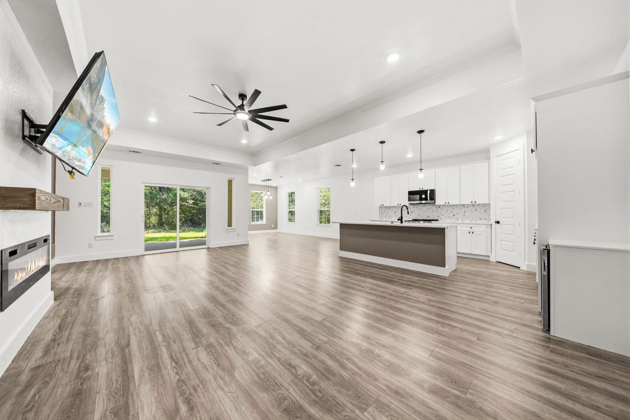 Spacious open-concept interior featuring wood-finish flooring, a contemporary kitchen island with pendant lighting, white cabinetry, and an electric fireplace with a wood mantel