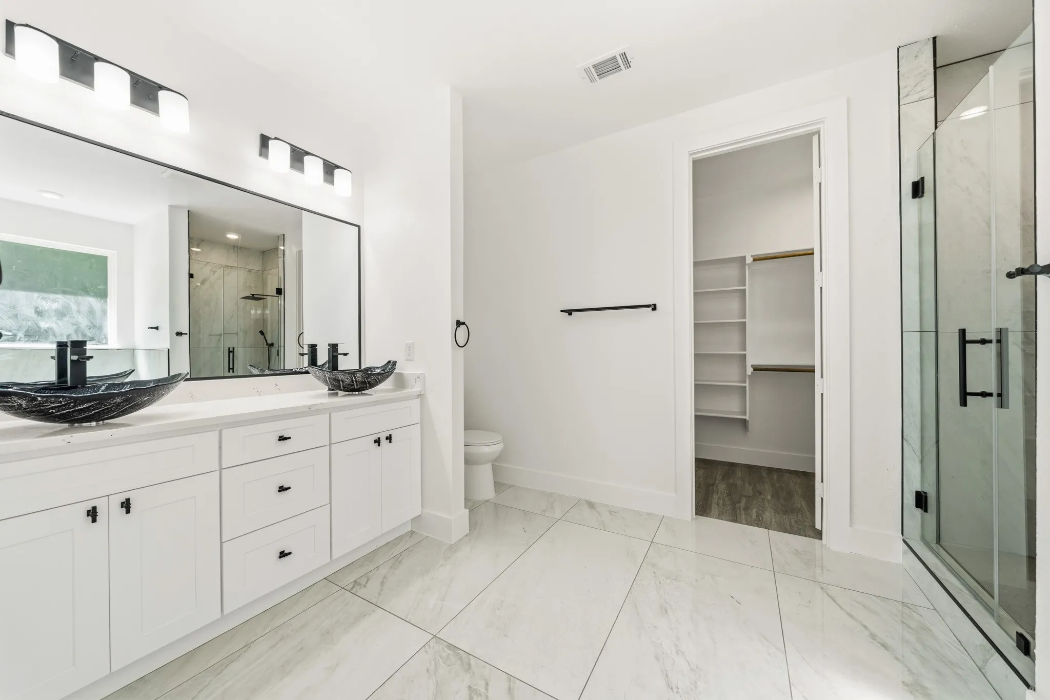 Spacious bathroom featuring a dual vanity with vessel sinks, a glass-enclosed shower, large format floor tiling, a separate water closet, and a walk-in closet with built-in shelving