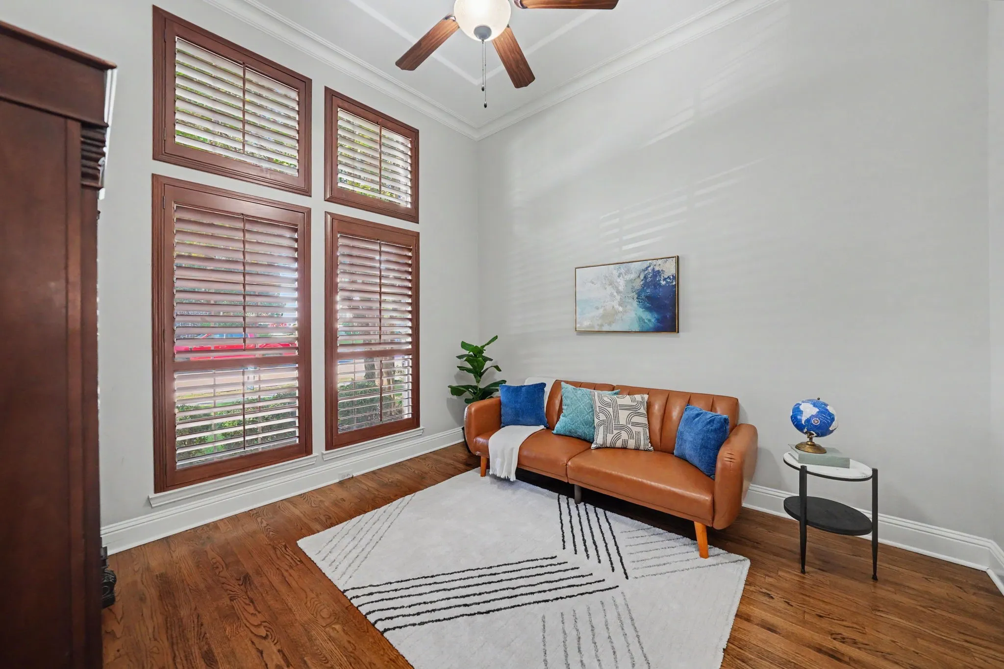 Hardwood flooring, crown molding, a ceiling fan, and tall windows with wood-frame plantation shutters
